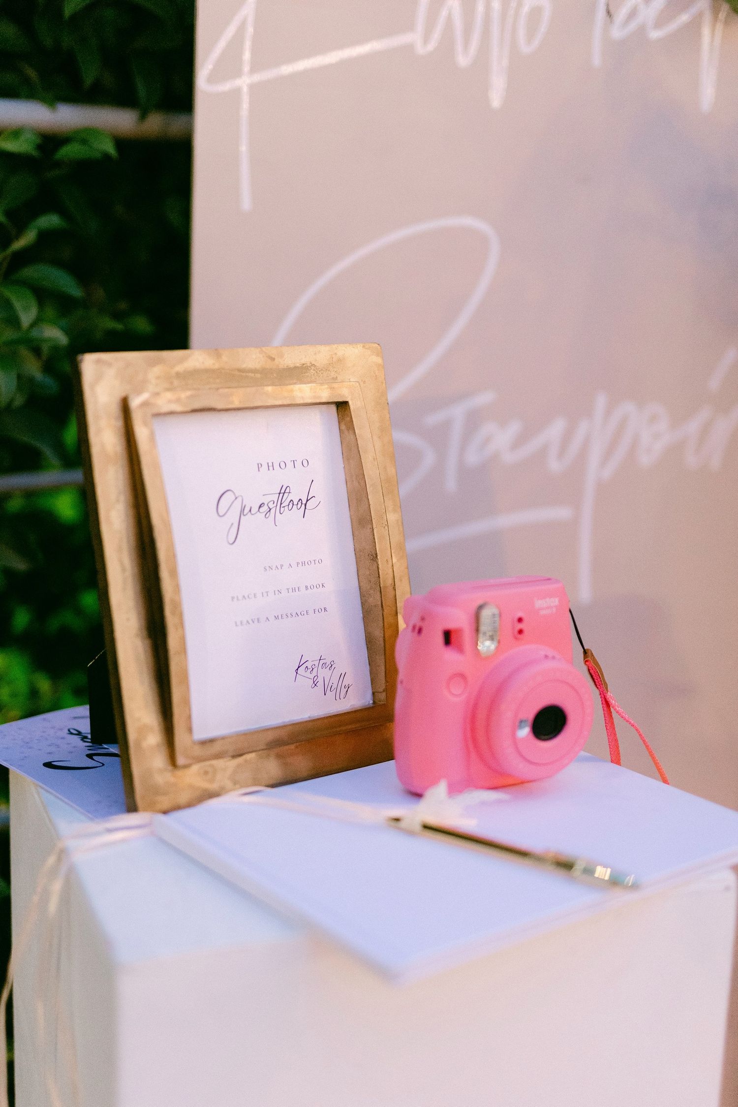 A pink Instax camera and guestbook setup on a white table with a wooden frame displaying wedding details.