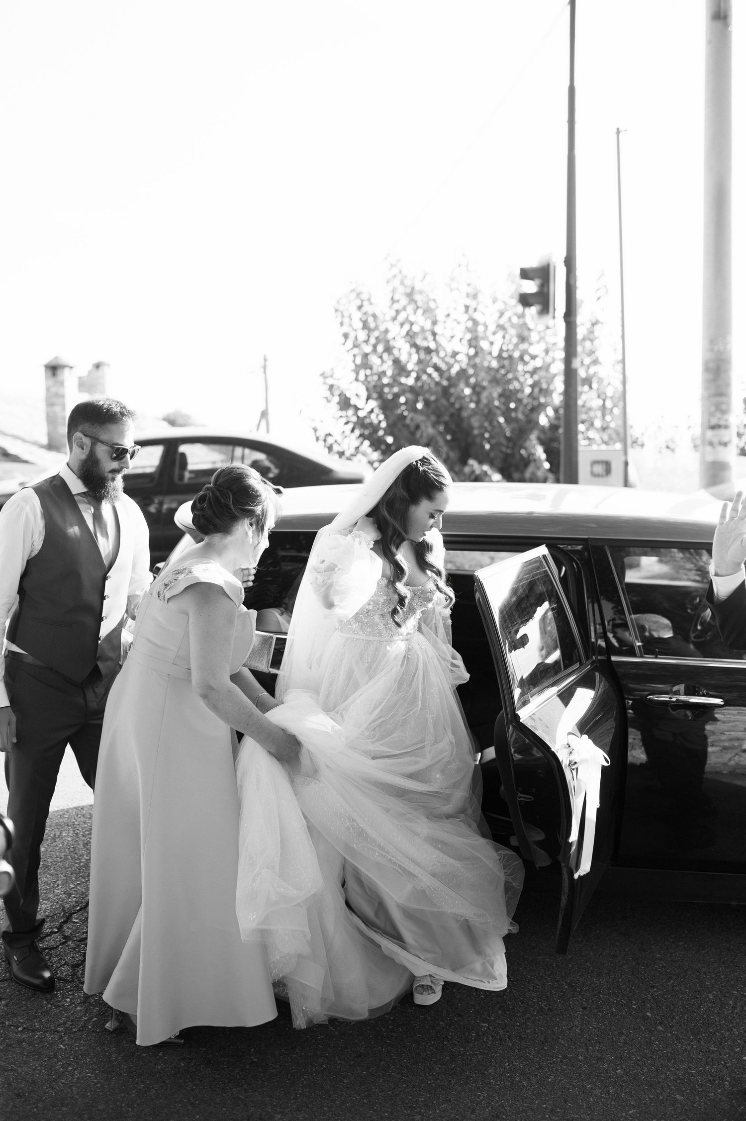 A bride in a flowing white wedding dress gets help entering a vintage car as attendants assist with her gown.