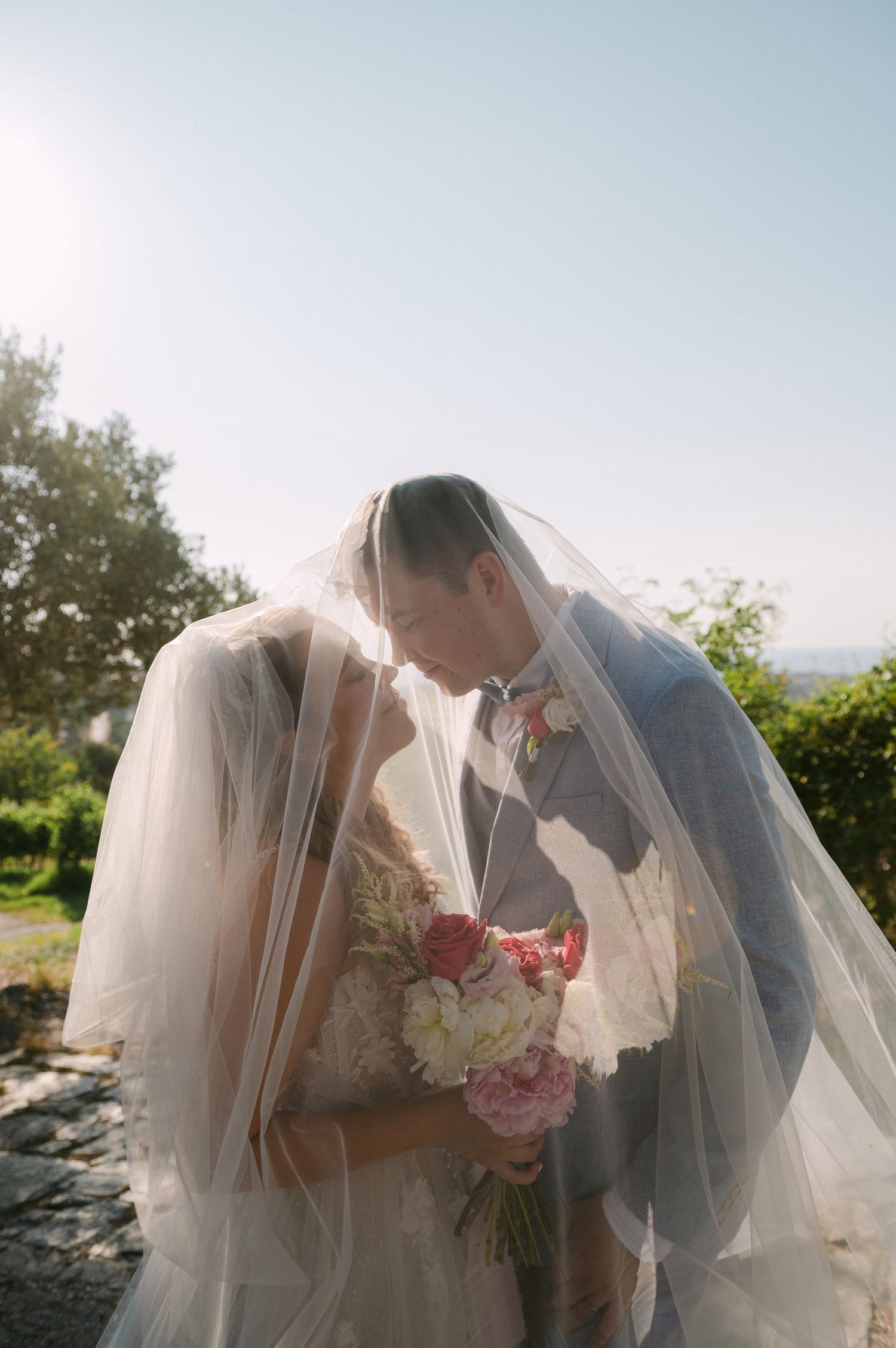 A romantic wedding moment captured outdoors with a flowing veil and pink floral bouquet against a scenic natural backdrop.
