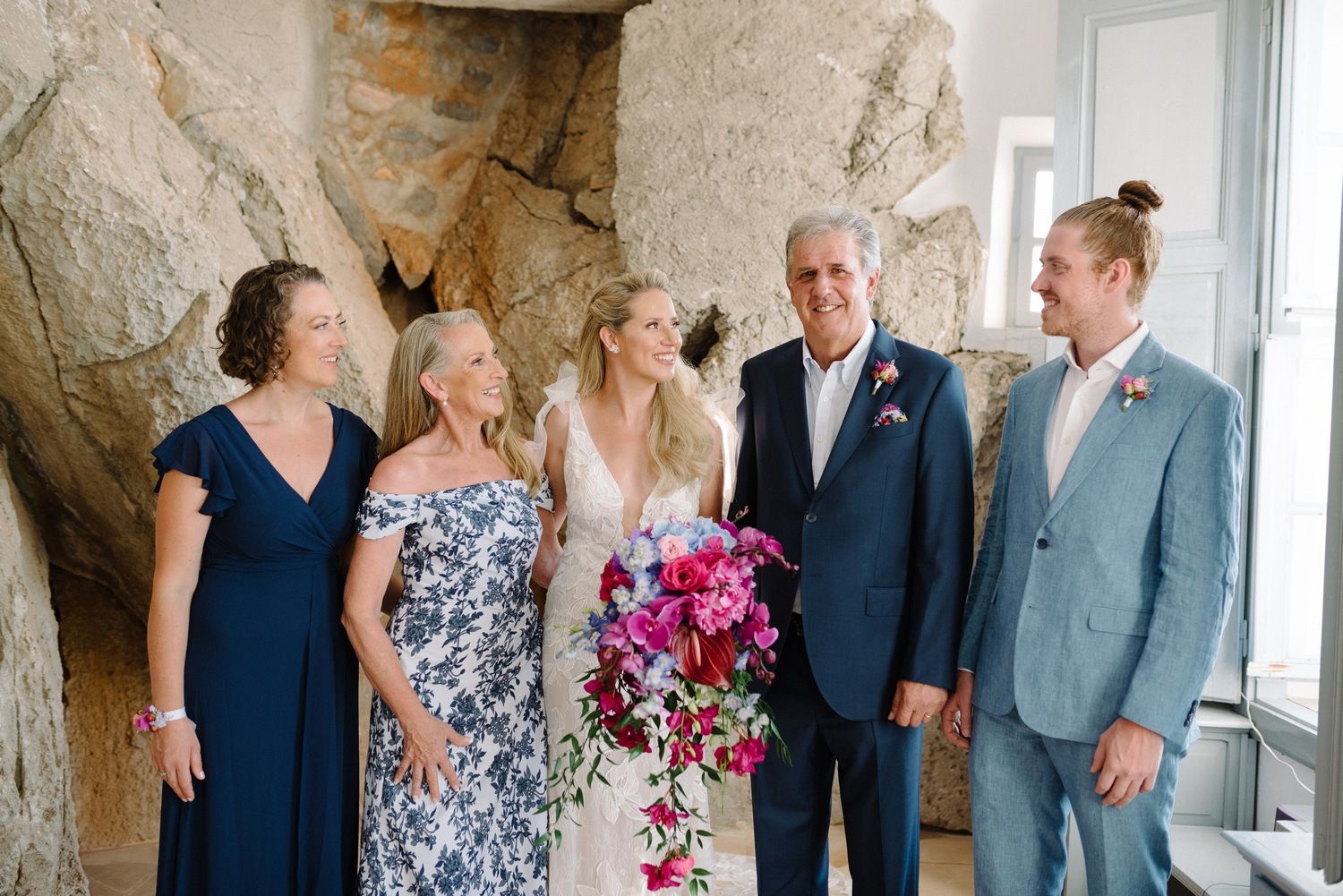 A wedding party poses together against a stone wall backdrop as they share joyful laughs.
