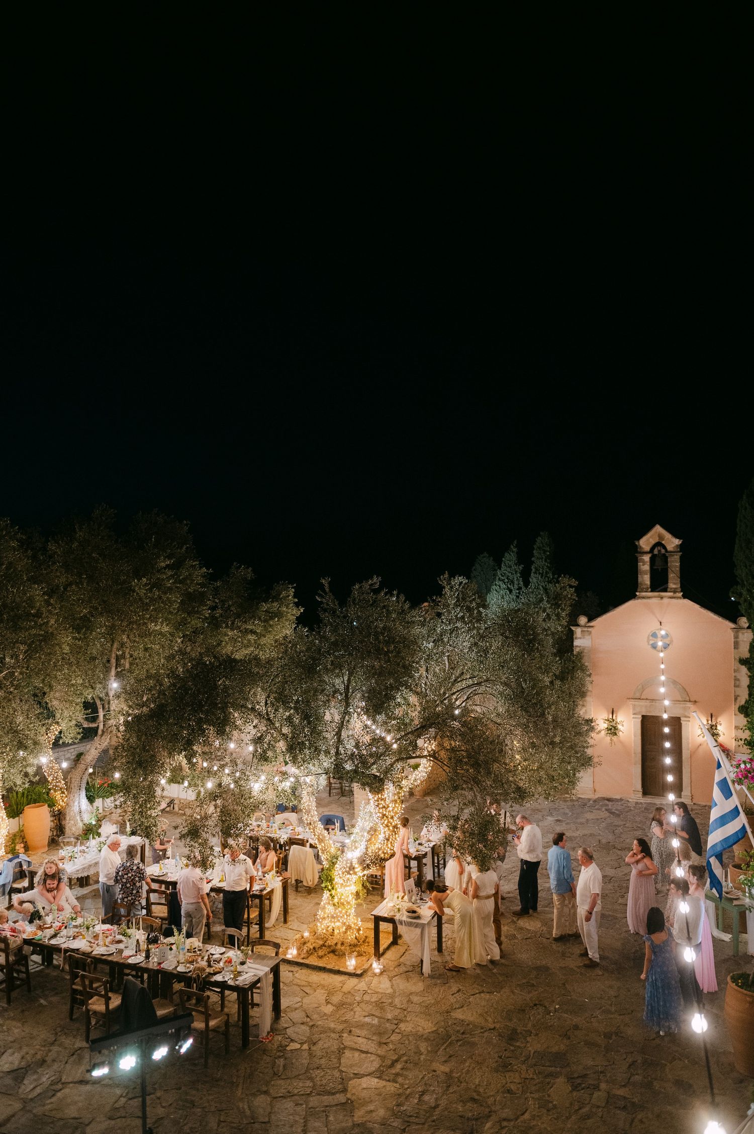 An outdoor evening wedding reception in Greece with twinkling lights and a small chapel under a dark night sky.