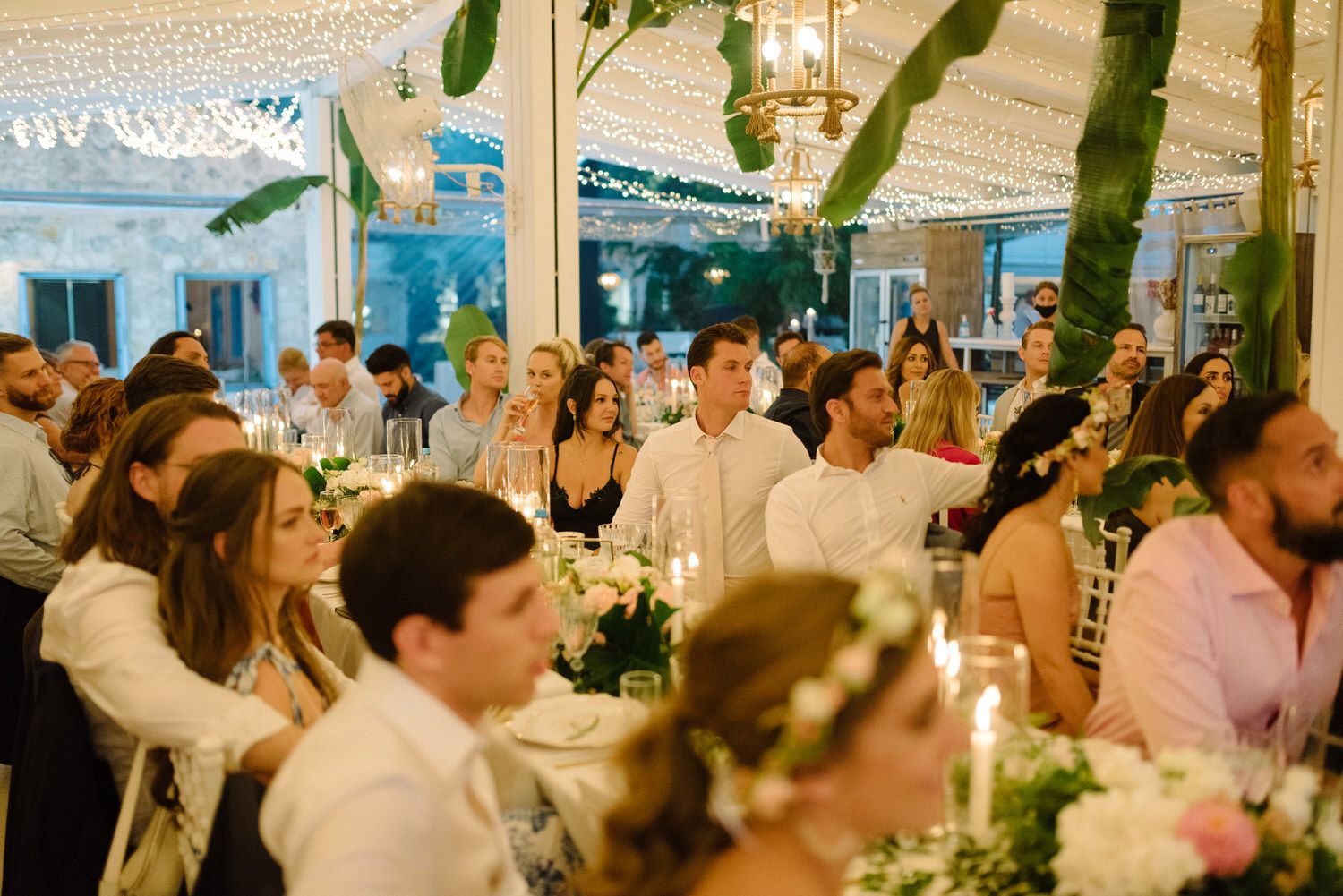 Wedding reception guests seated at elegant tables under a white tent with twinkling lights and greenery.