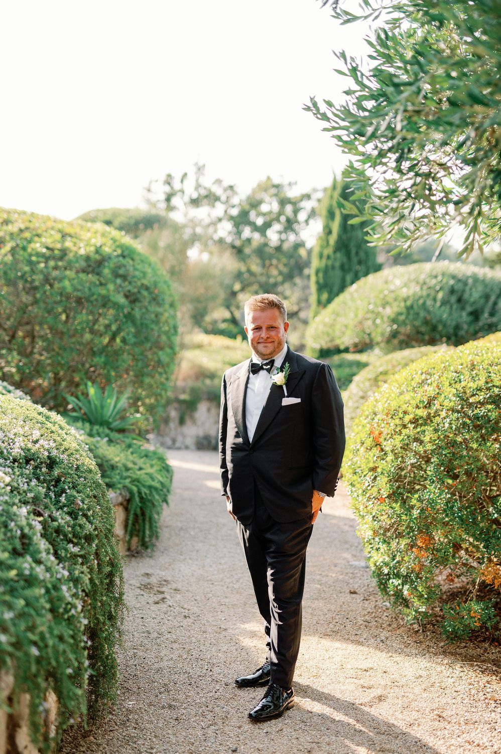 Groom in classic black tuxedo poses on garden pathway with cypress trees.