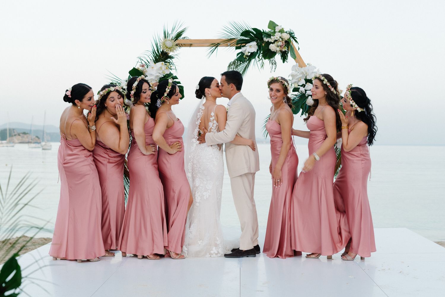 A wedding party poses under an arch with bridesmaids in pink dresses on a white platform.