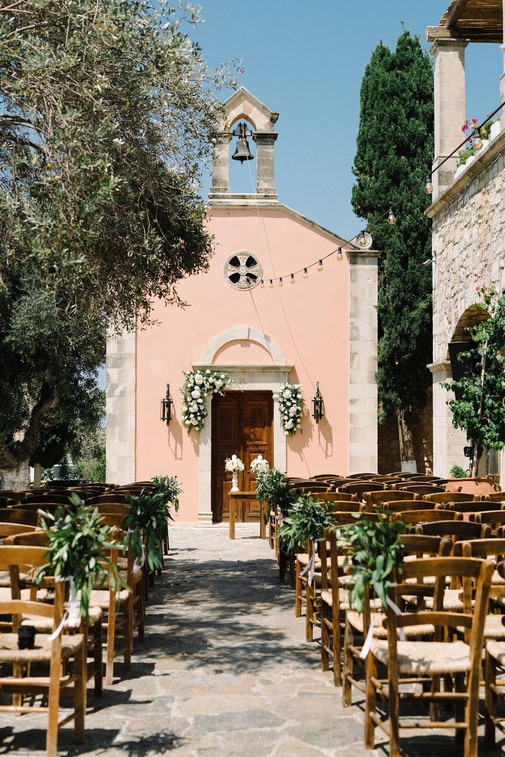 Pink chapel with wooden chairs set up for outdoor wedding ceremony under olive trees.
