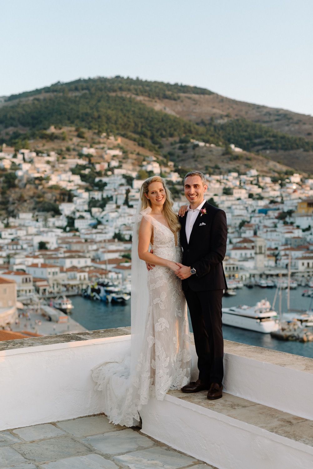Newlyweds pose on white terrace overlooking Mediterranean coastal town and harbor with hillside in background.