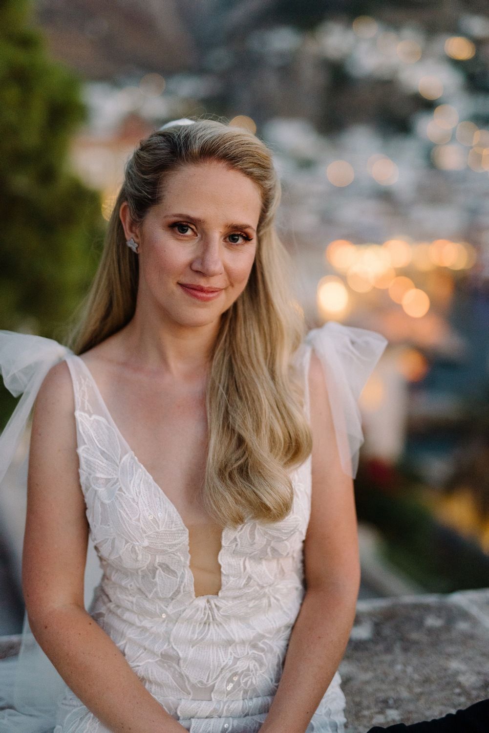 Bride in lace V-neck gown poses against twilight coastal backdrop.