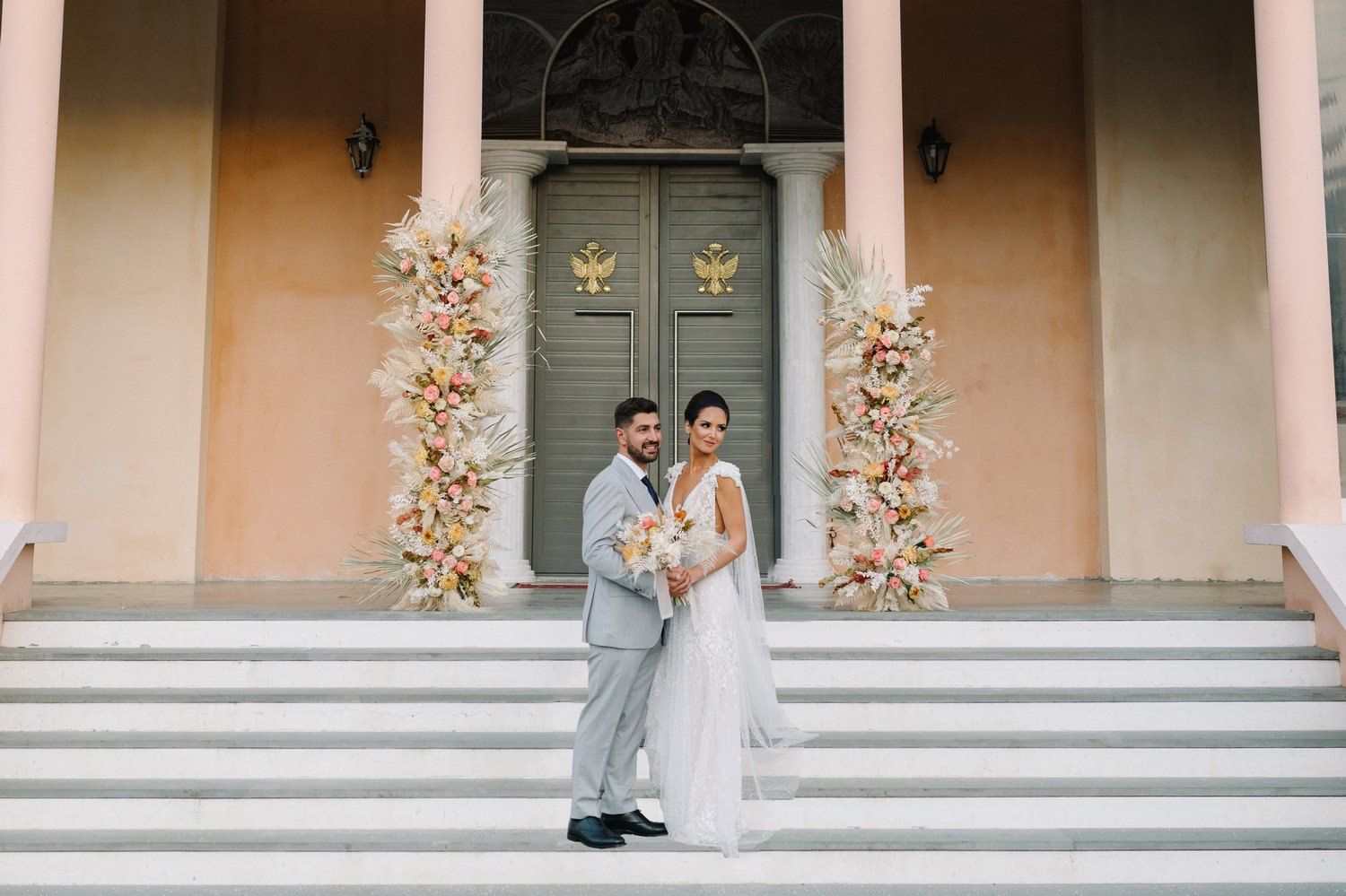 Elegant couple standing on grand steps in front of ornate floral-decorated doorway.