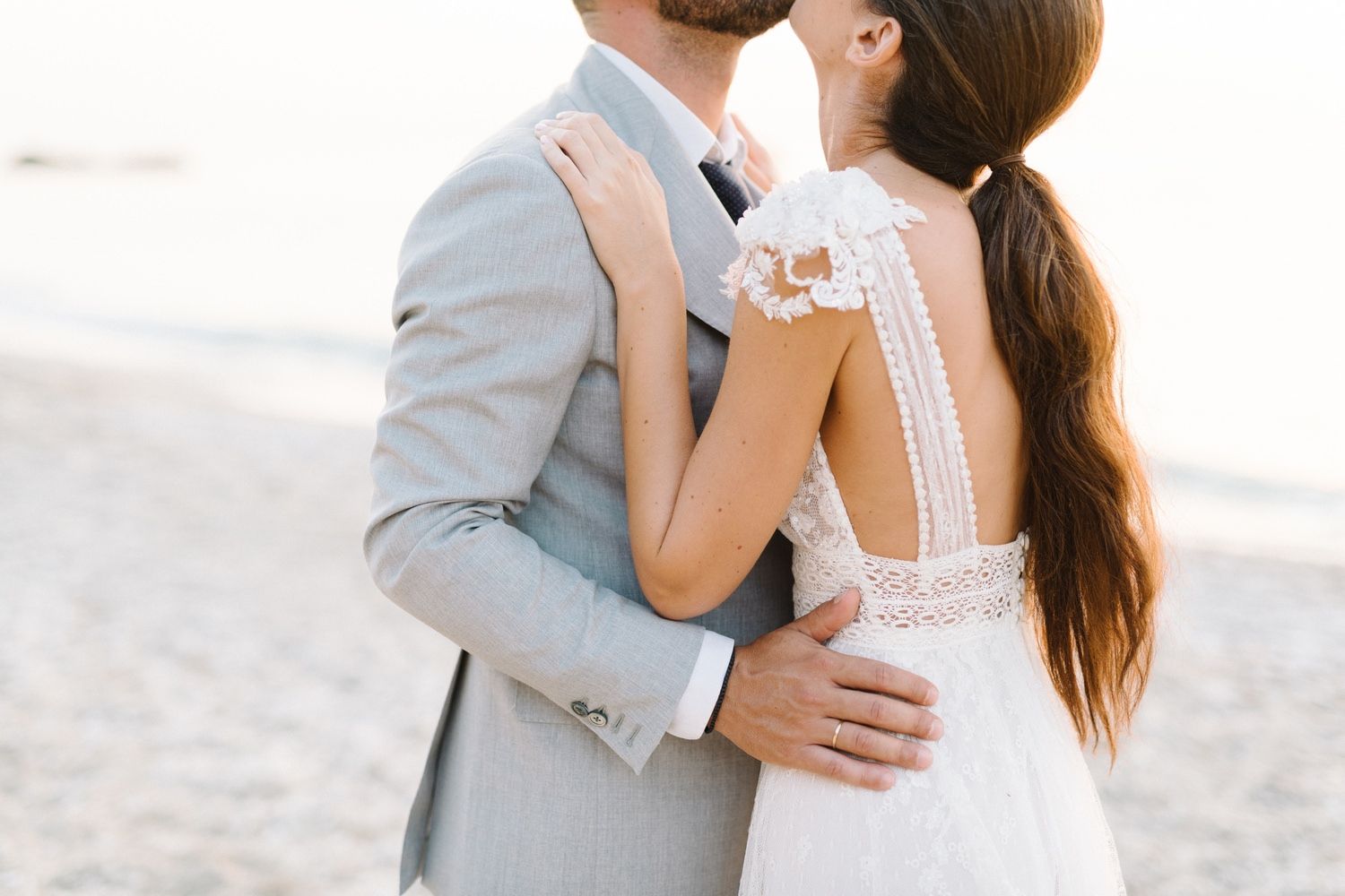 A close-up wedding photo highlighting the detailed back of a white lace dress against a light grey suit.