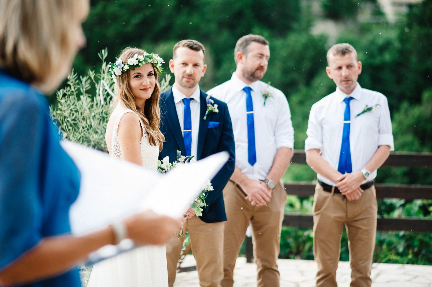 Wedding party stands outdoors with groomsmen in khaki pants and blue ties during summer ceremony.