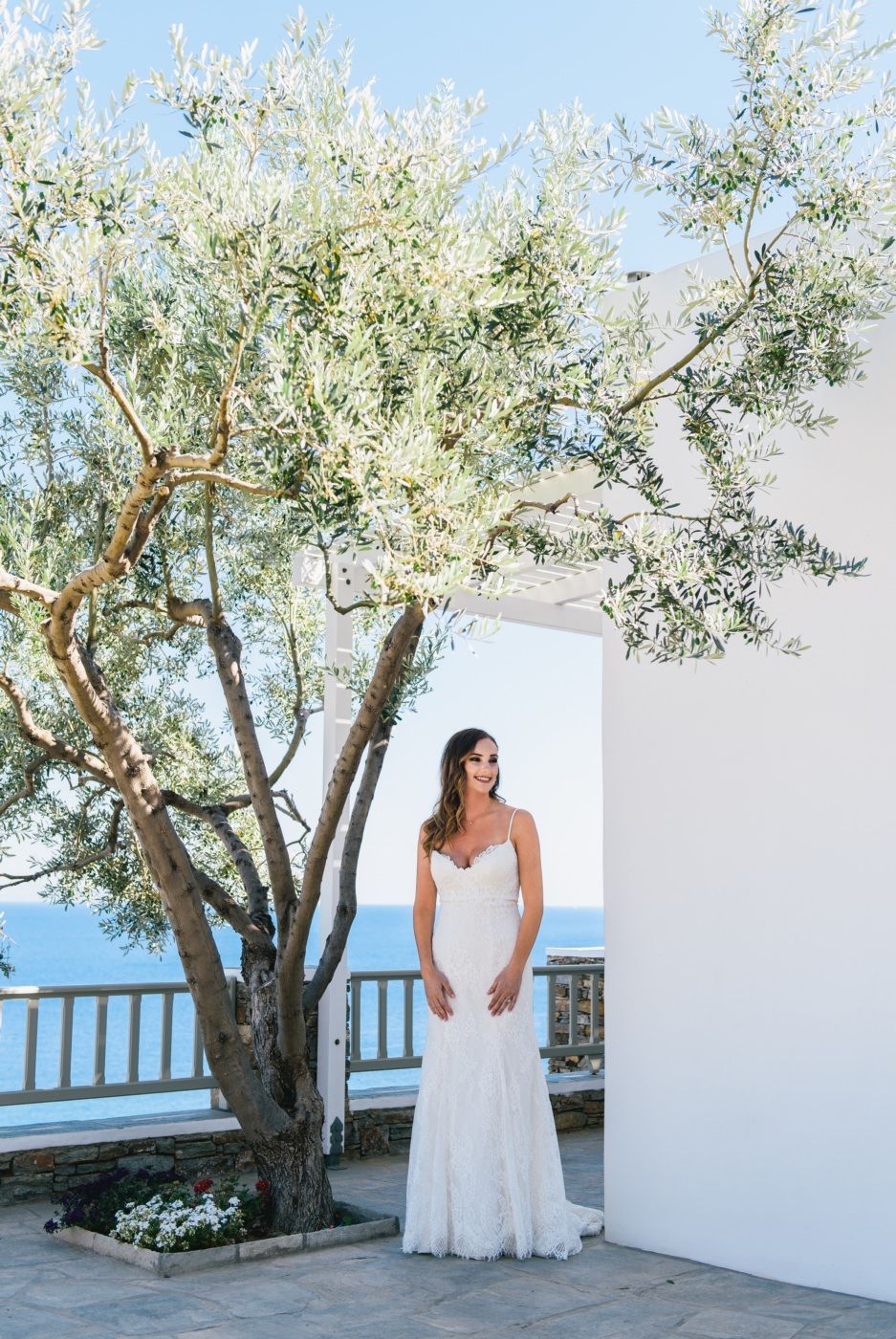 White wedding dress against Mediterranean architecture with olive tree and ocean view.