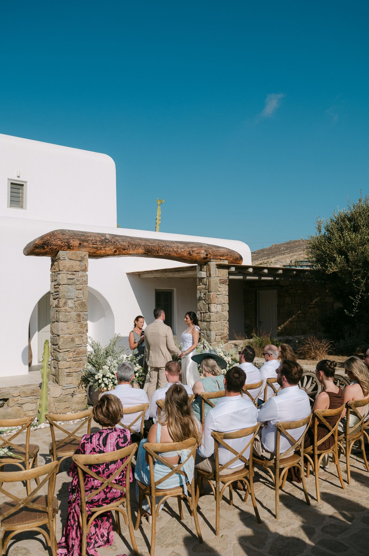 Outdoor wedding ceremony setup with wooden cross-back chairs arranged in rows against a white Mediterranean-style building.