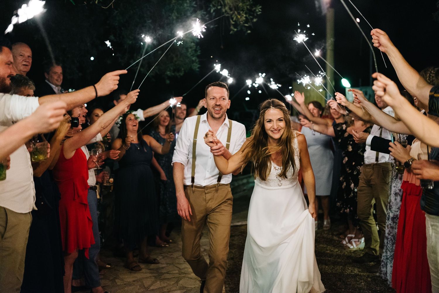 Wedding couple exits through sparkler tunnel held by guests in red dresses at nighttime celebration.
