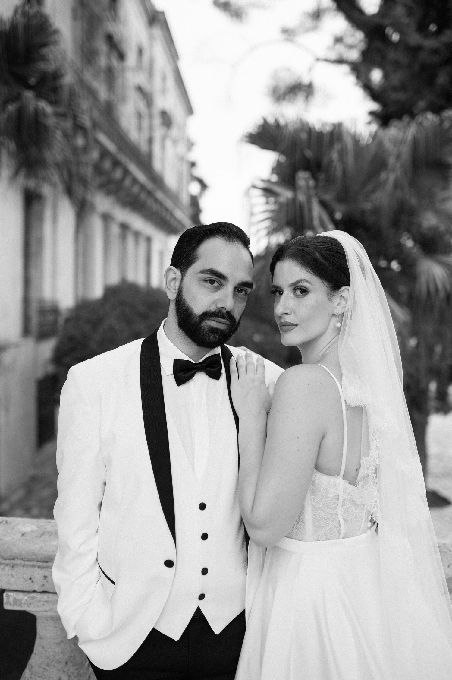 Elegant black and white wedding photo of a couple in white formal attire posing together outdoors.