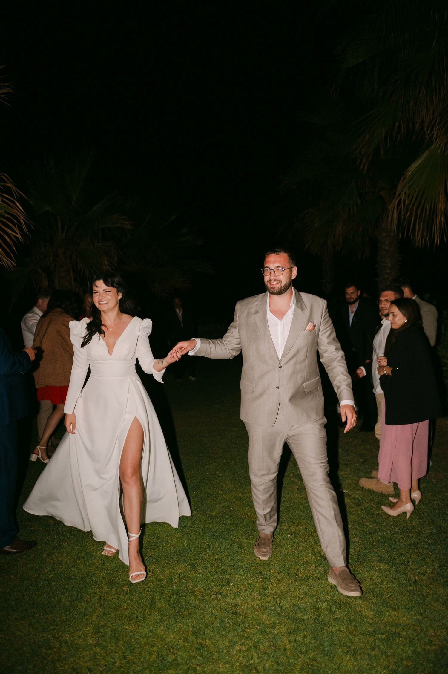 A couple dances together on a grassy lawn at night, both wearing elegant white and beige formal attire.