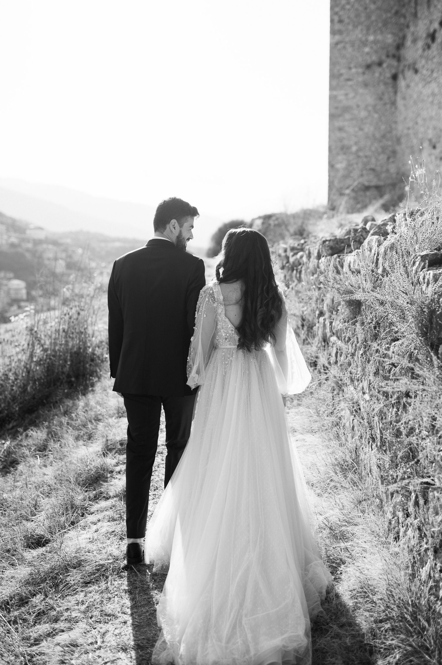 A couple in wedding attire walking along a scenic mountain path in black and white photography.