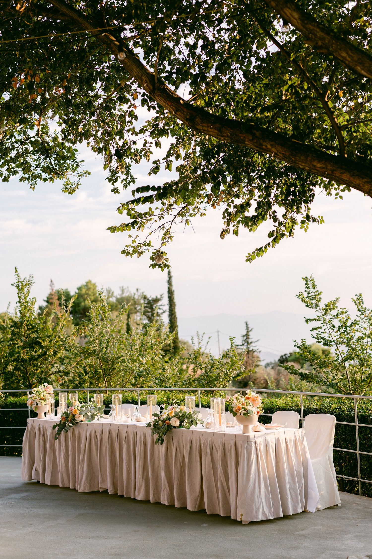 An elegant outdoor wedding reception table with blush tablecloths set under a tree canopy on a terrace.