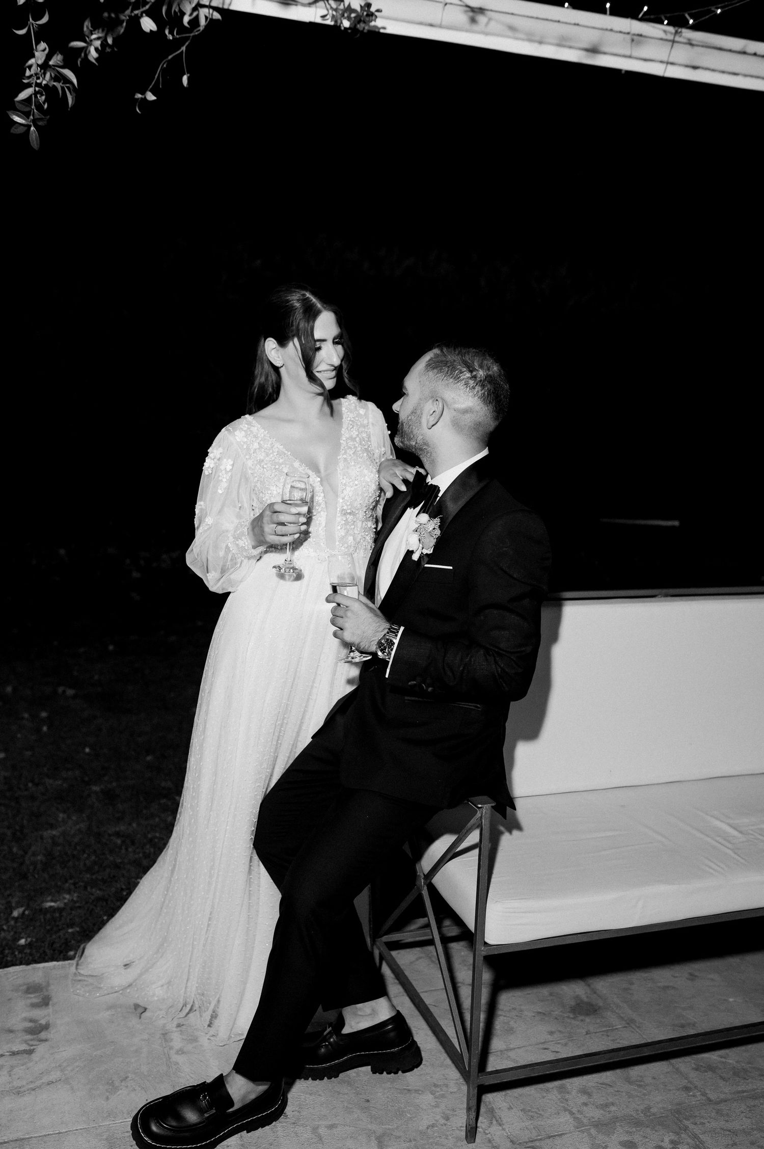 A couple in wedding attire shares an intimate moment on a bench at night in black and white.