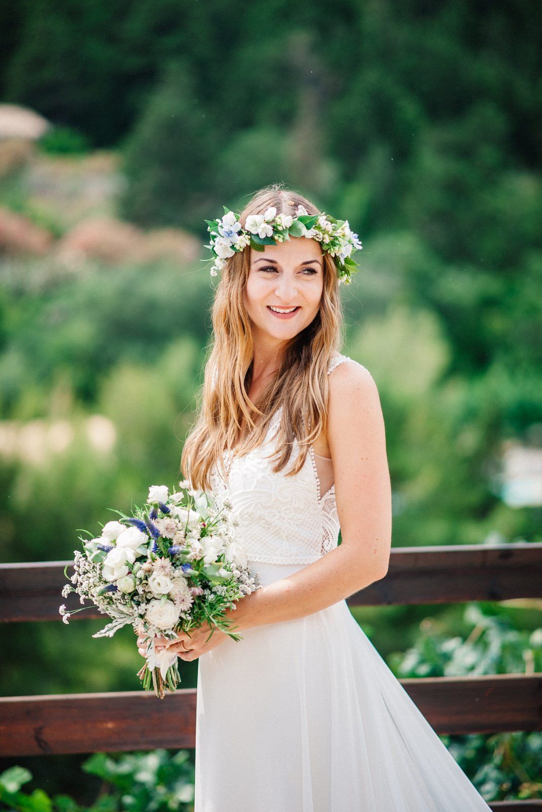 Bride in white dress with floral crown and bouquet poses against mountain landscape backdrop.