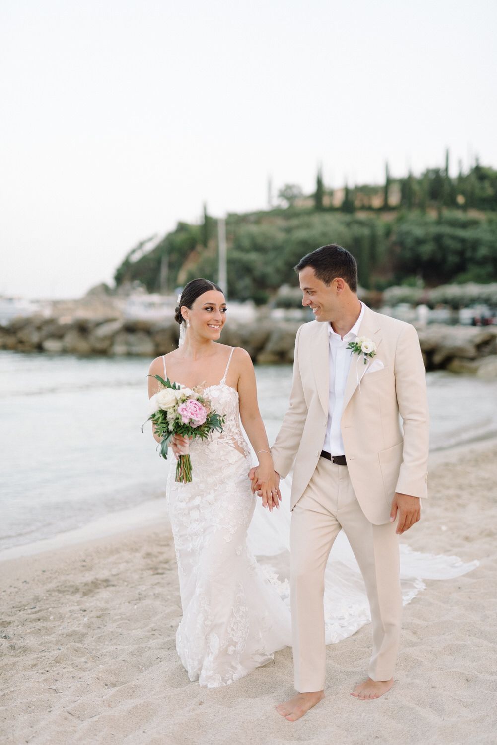 Couple walking barefoot on beach with coastal cliffs in background.