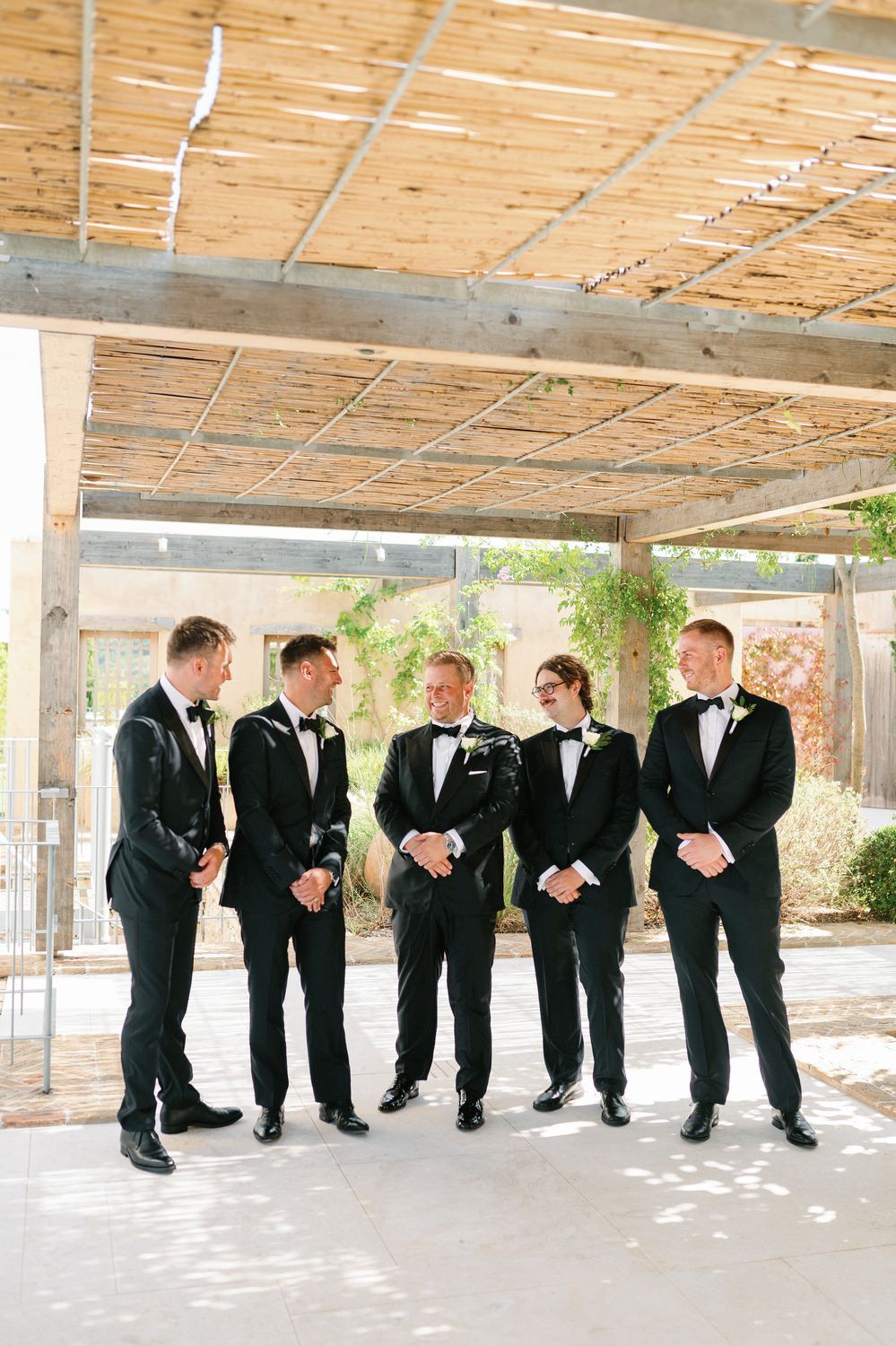 A group of groomsmen in black tuxedos and bow ties share a laugh while standing under a wooden pergola.