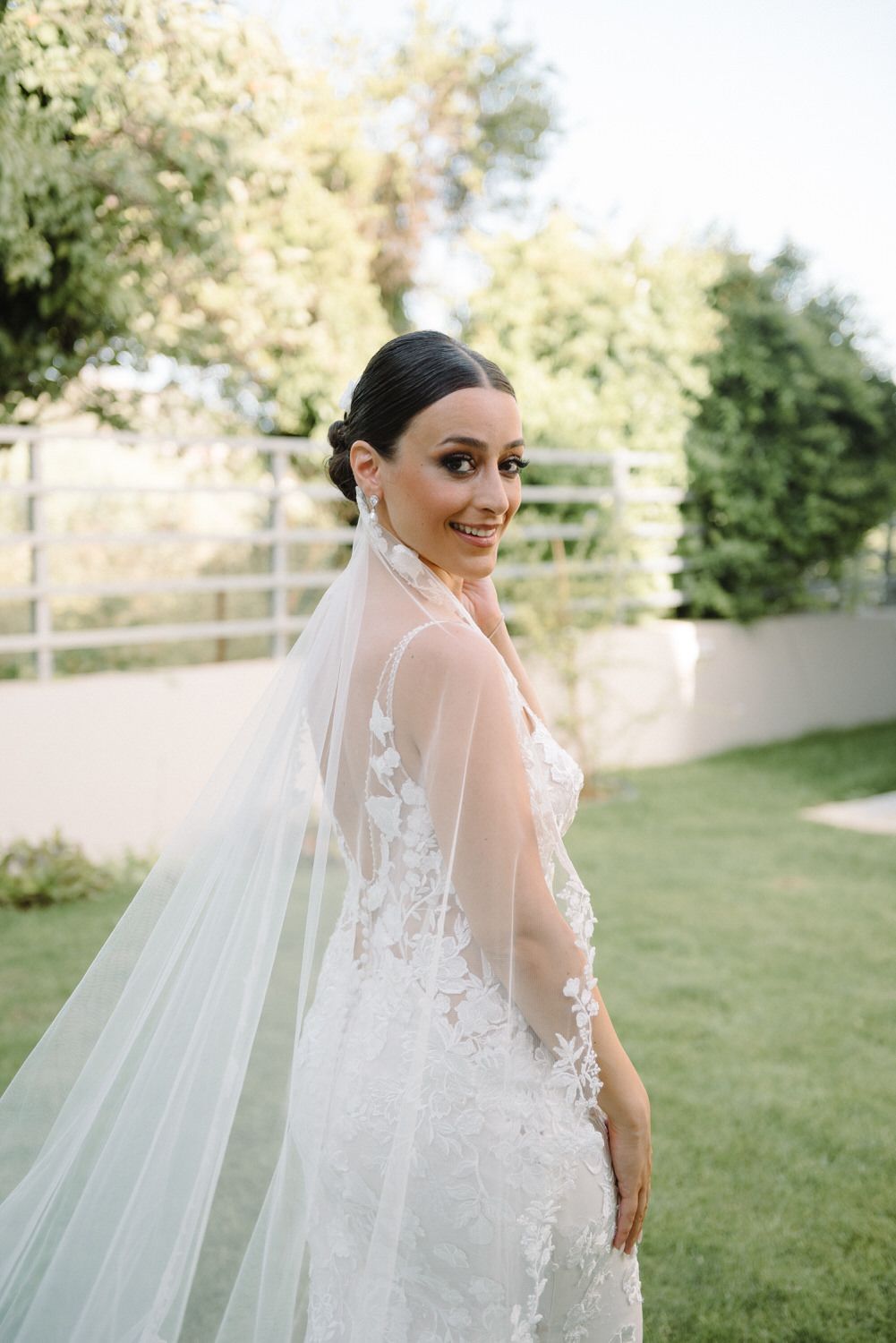 Bride in fitted lace gown with flowing veil smiles over shoulder in garden setting.