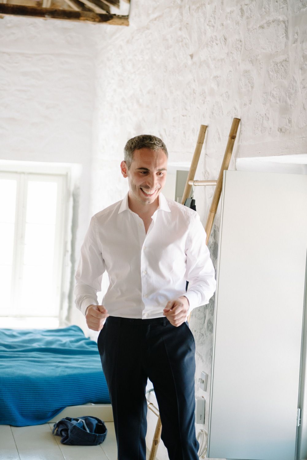 A man in a crisp white shirt and dark pants stands confidently in a bright room.
