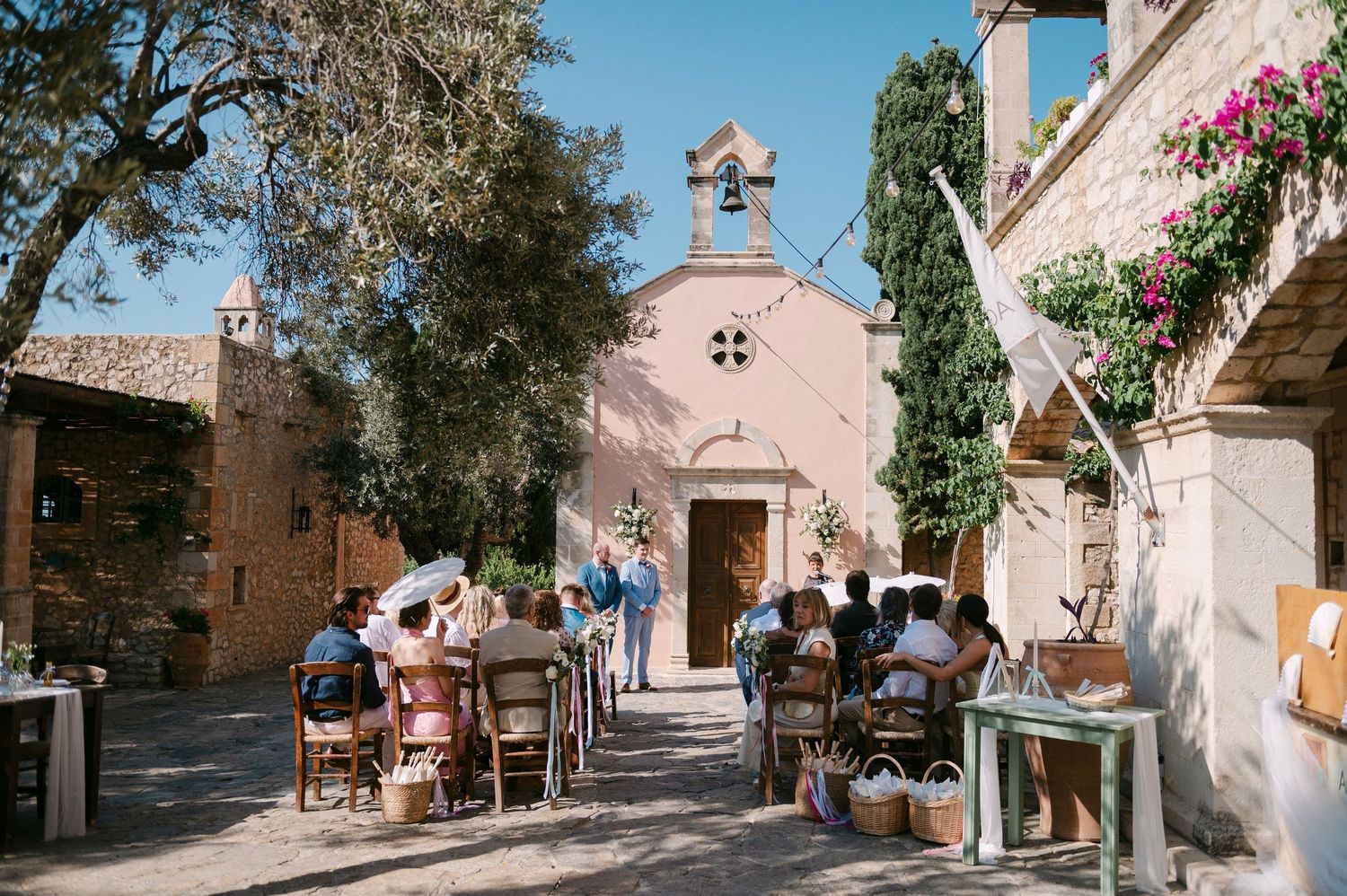 Sun-drenched Mediterranean street with pink walls, flowering trees, and cobblestone paths in a scenic Greek village.