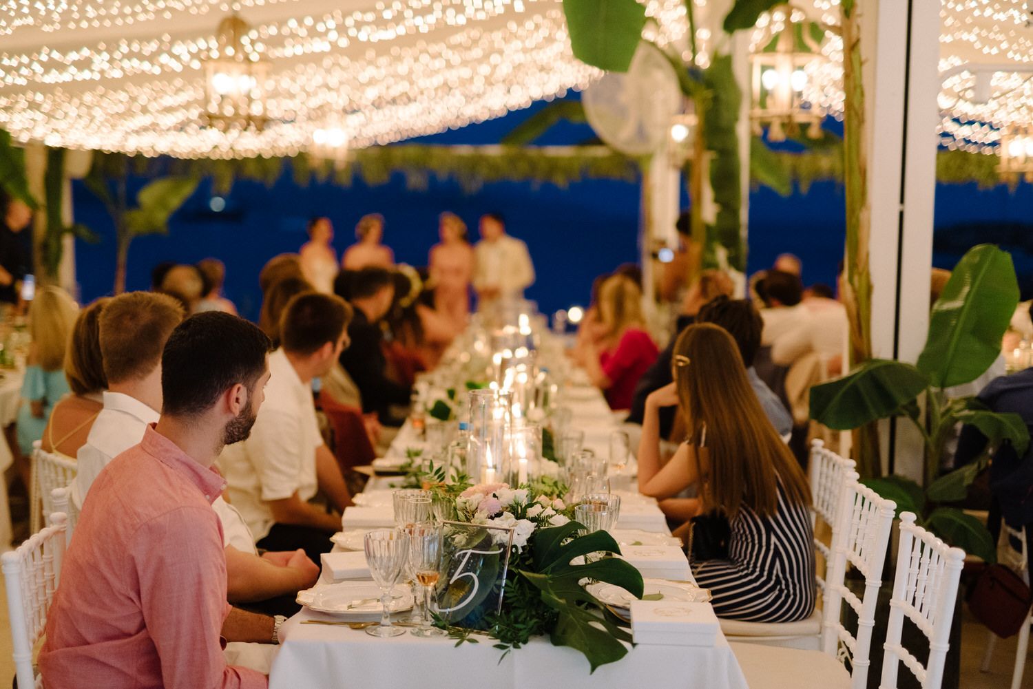 Long dining table set for an elegant outdoor wedding reception under string lights and greenery.