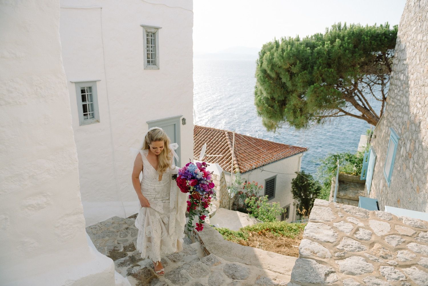 A person in white dress walks up Mediterranean stone steps with ocean view in background.