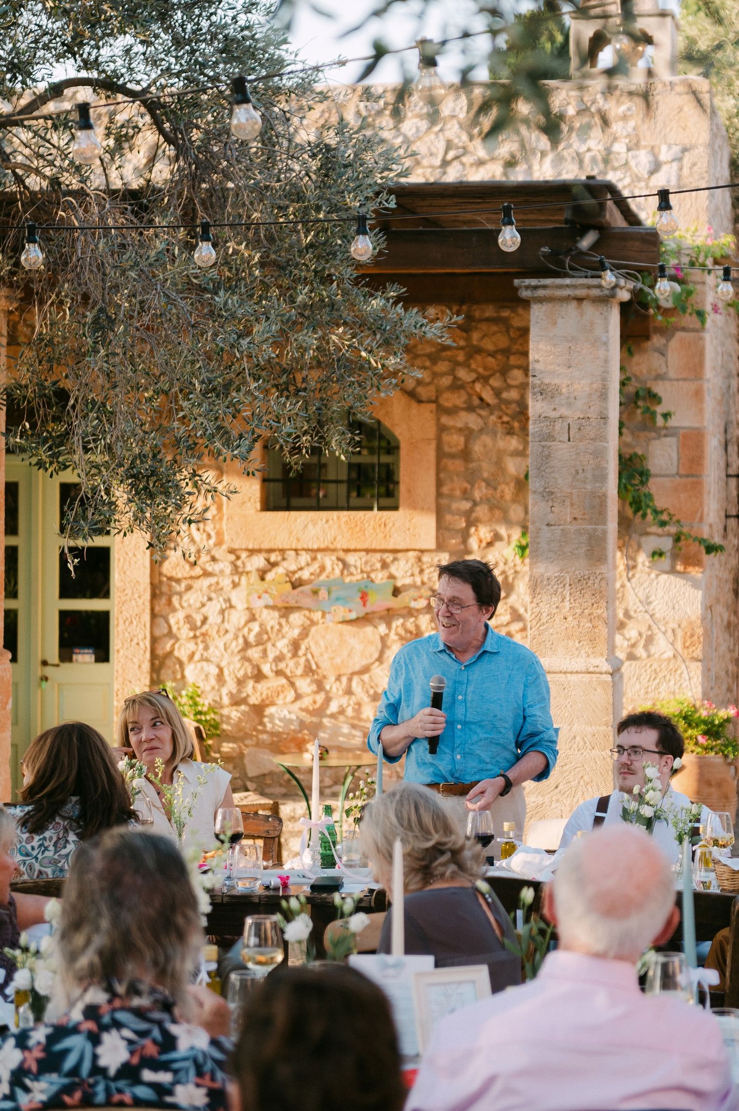 Wedding guests gather at an outdoor venue with stone walls and string lights creating a warm ambiance at dusk.