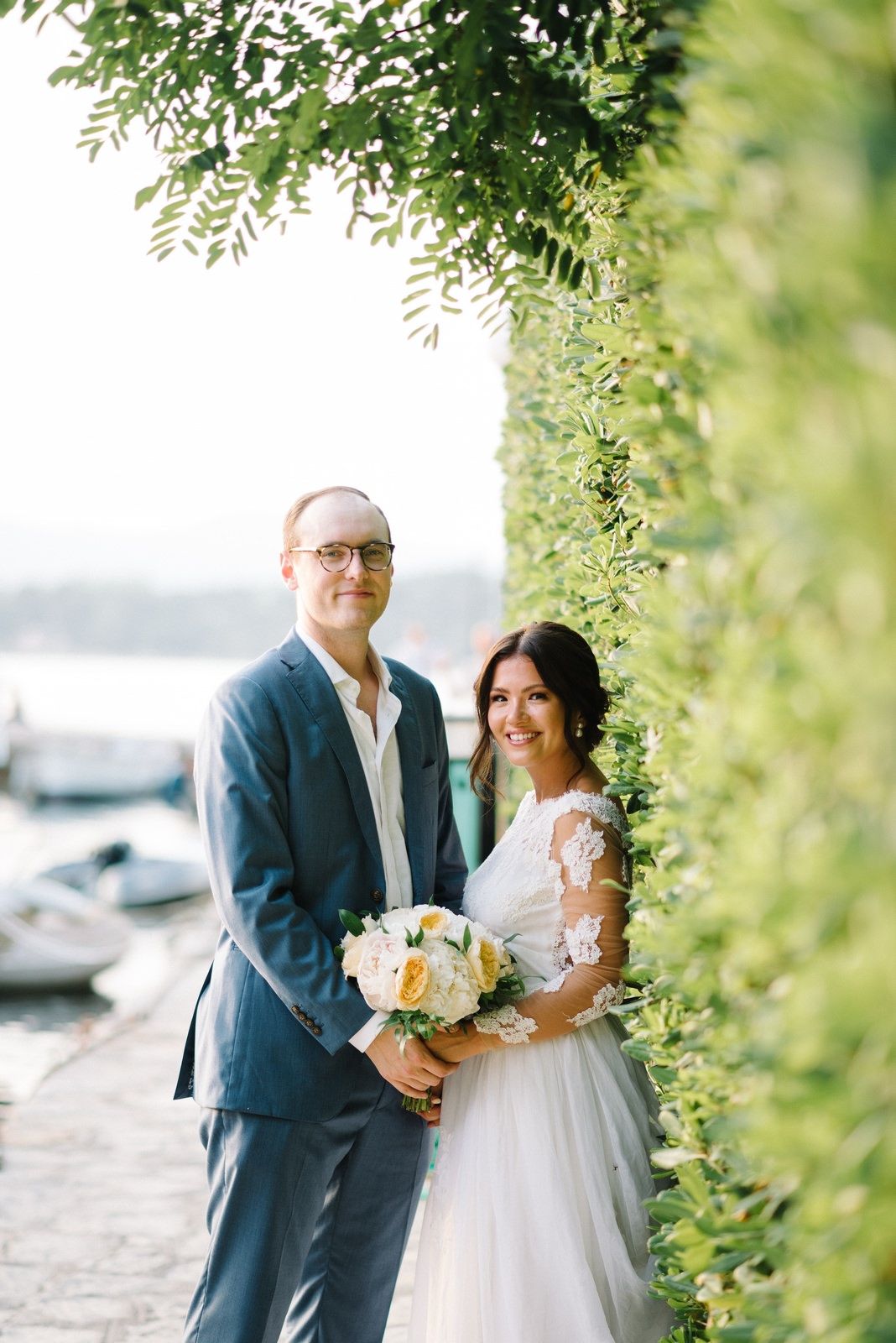 A wedding couple poses by a marina with lush greenery overhead and boats in the background.