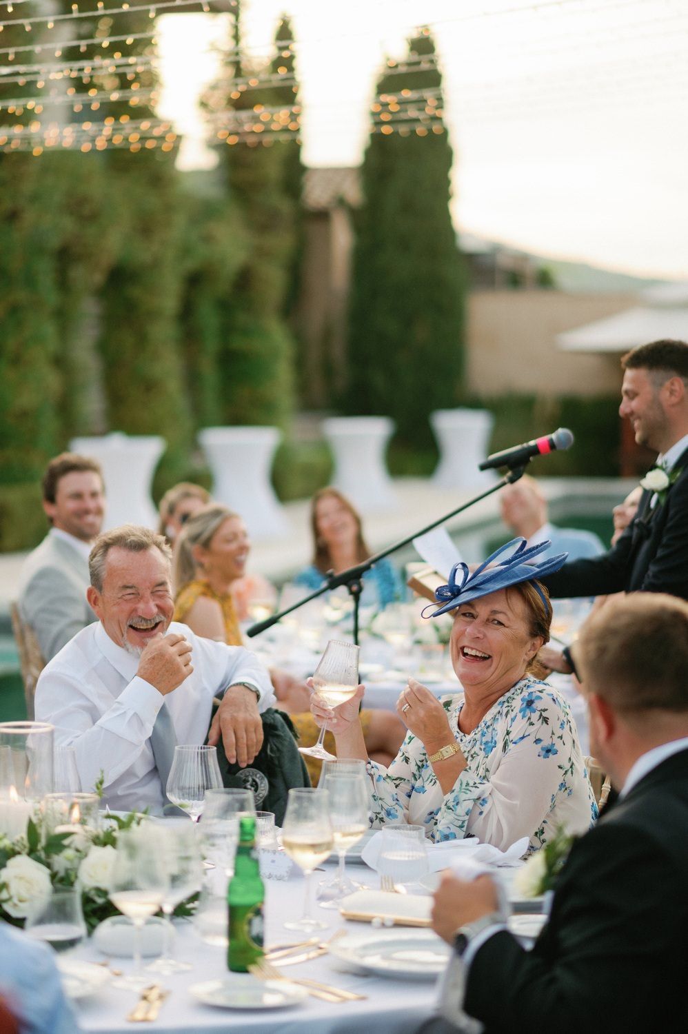 Guests laugh during speeches at an outdoor wedding reception under string lights.