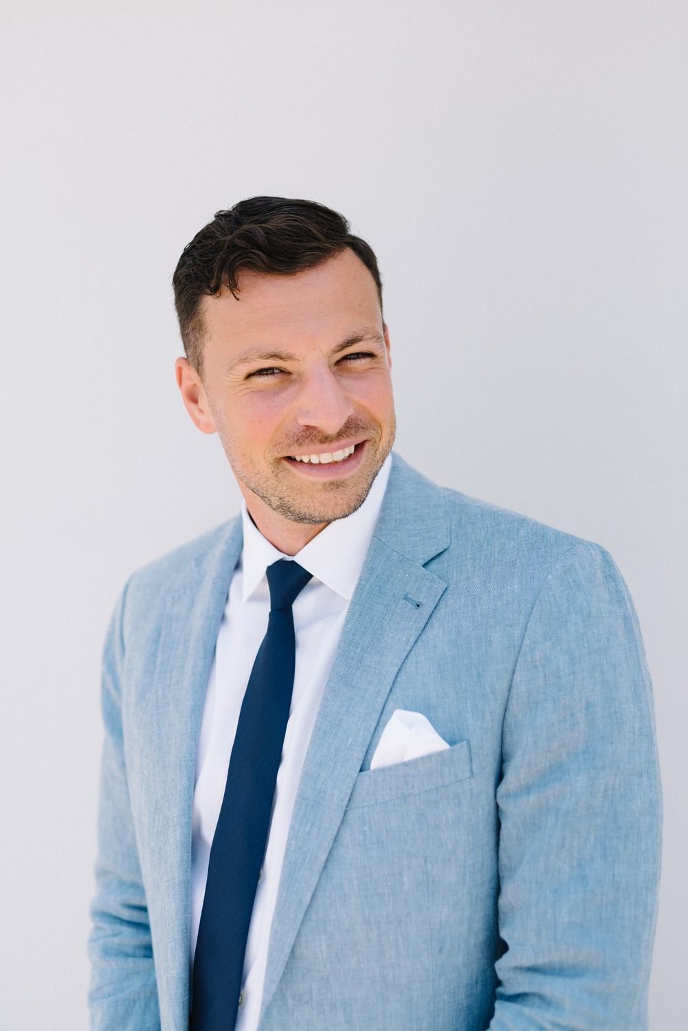 Man in light blue suit and navy tie smiling against white background.