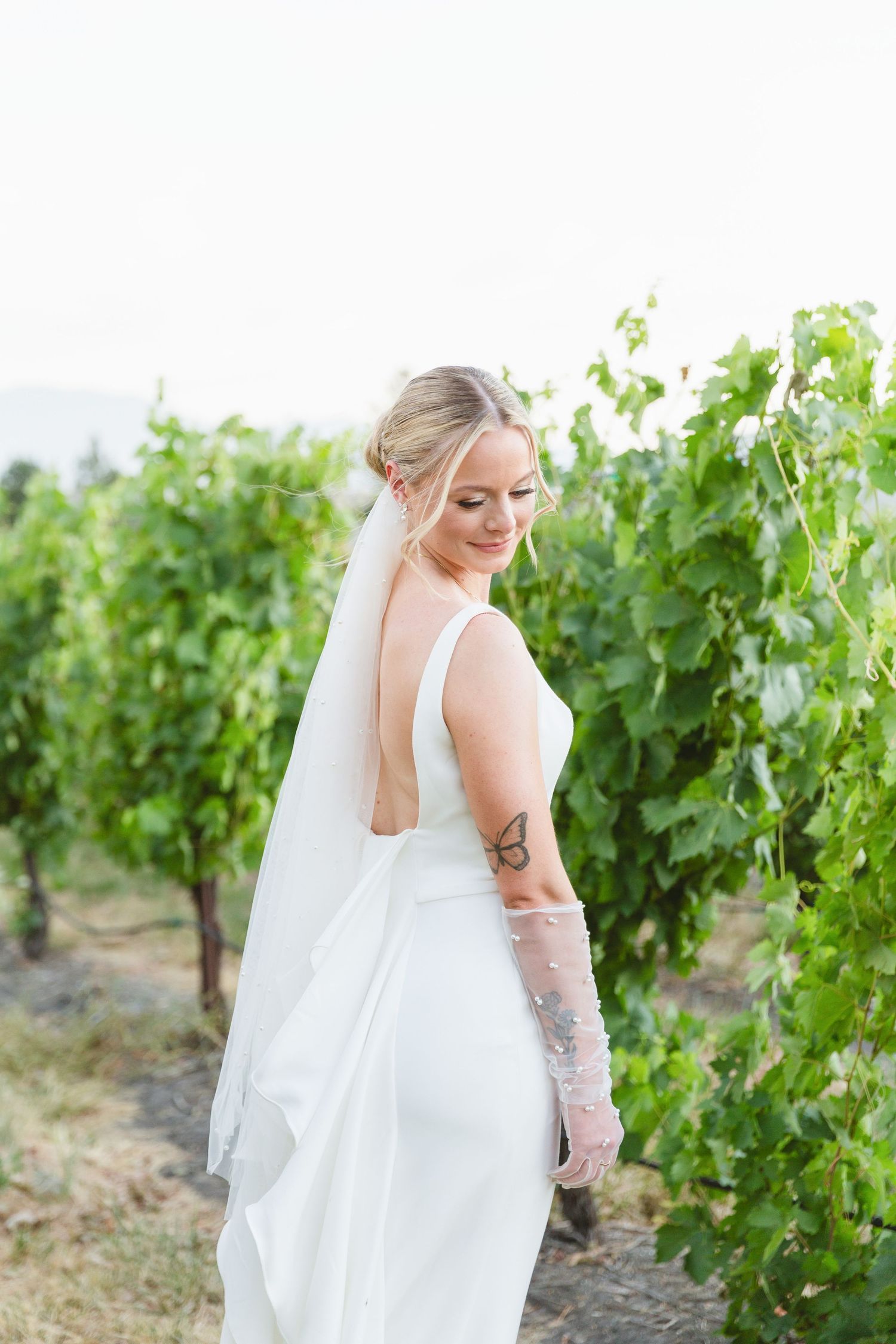 A bride in a white wedding dress poses in front of vineyard rows on a sunny day.