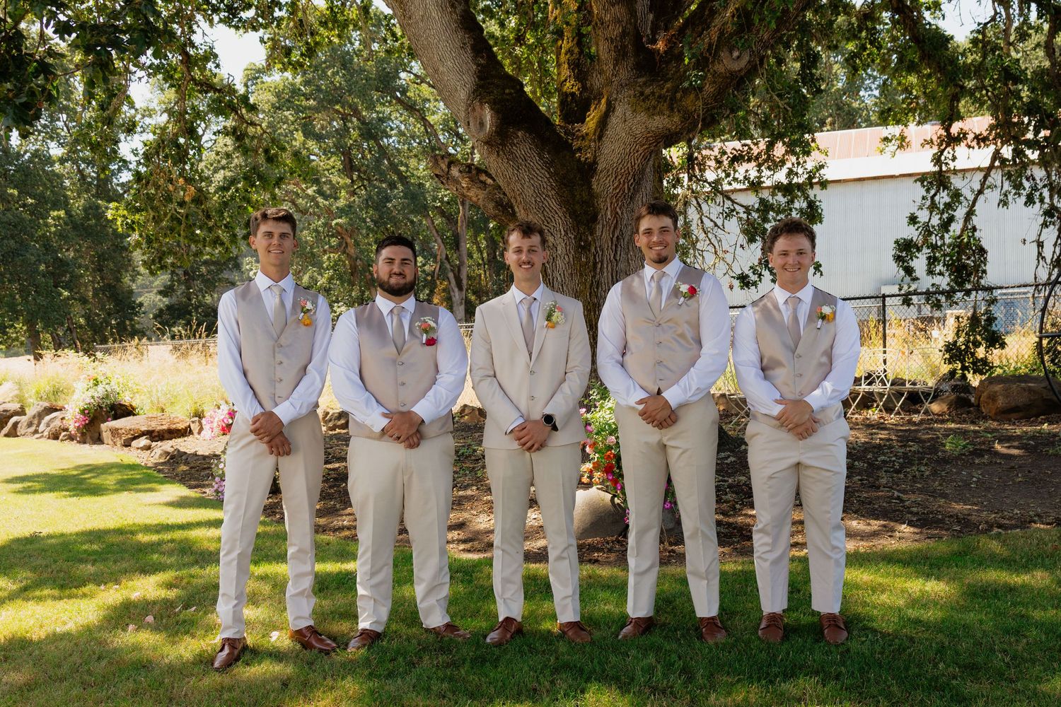 Five groomsmen in matching beige suits with floral boutonnieres posing under large tree outdoors.