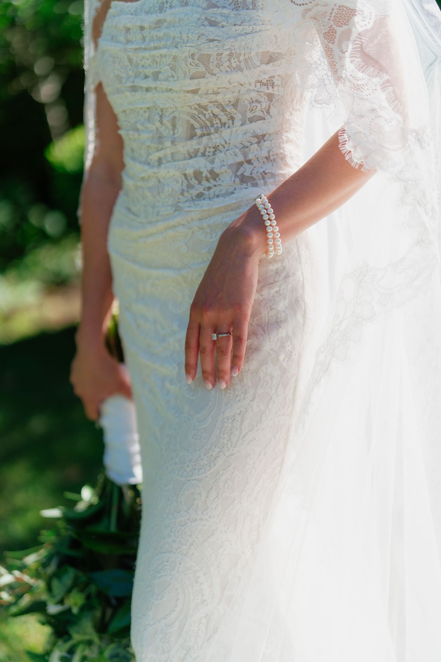 Close-up of bride in lace wedding dress showing pearl bracelet and engagement ring while holding bouquet outdoors.