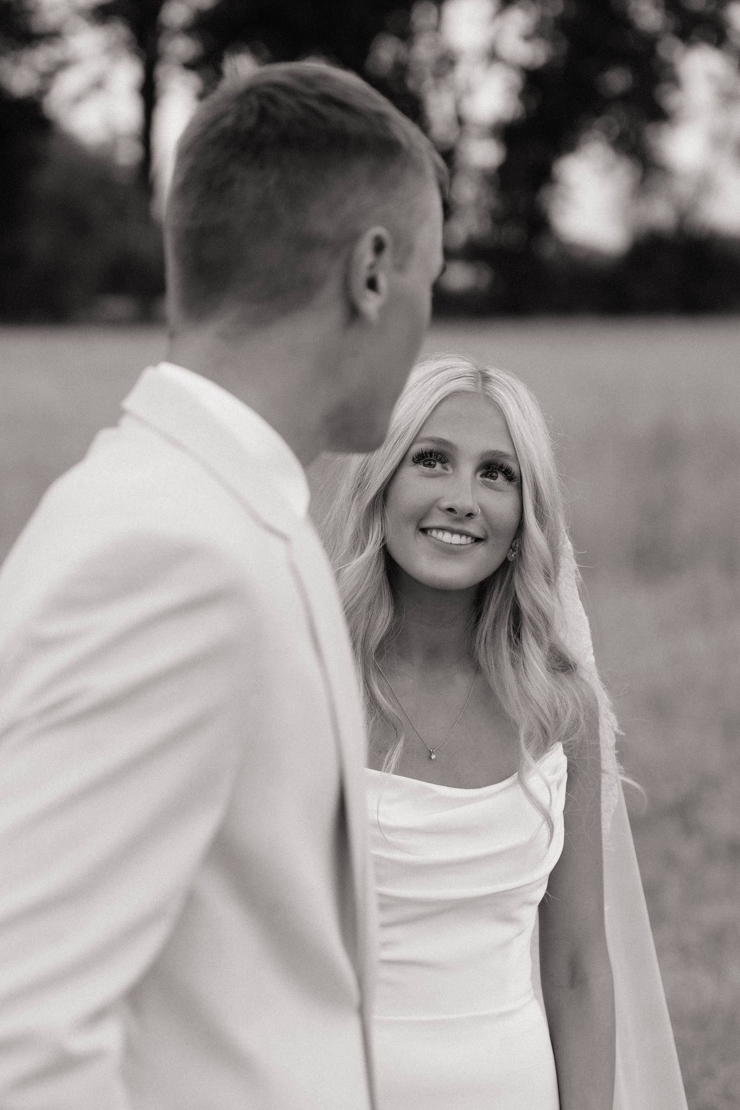 Black and white wedding photo of smiling blonde bride in white gown looking up at groom in white suit outdoors.