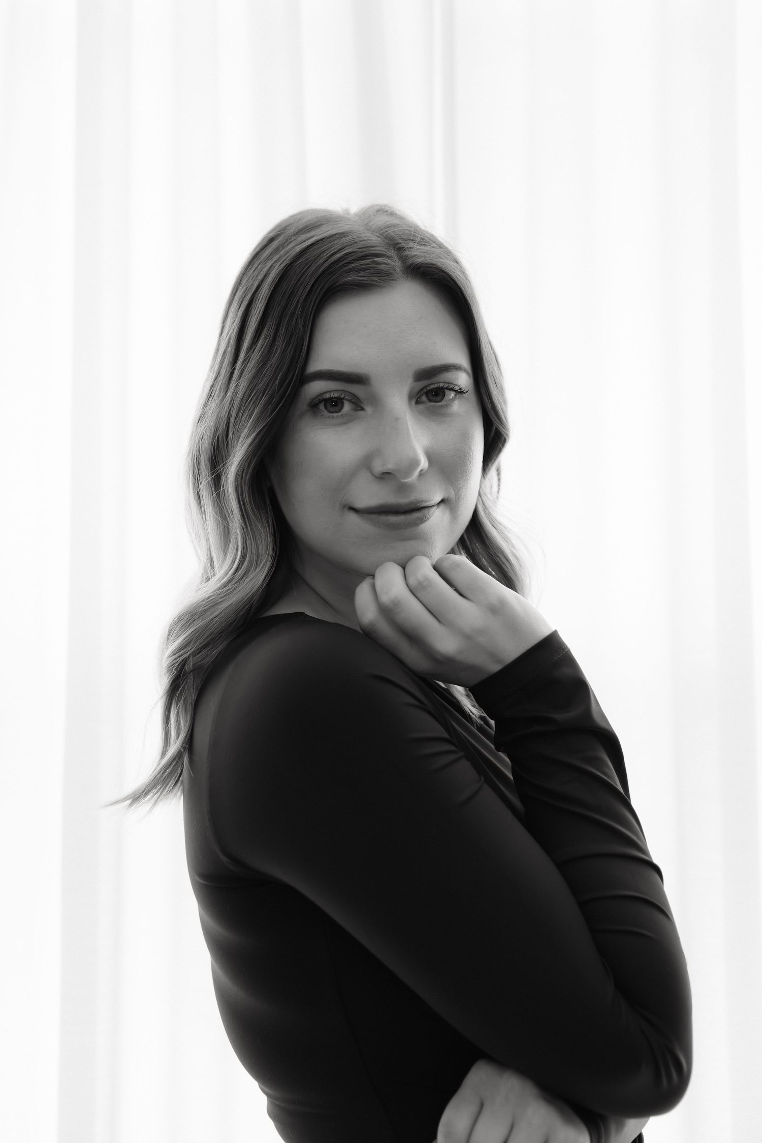 Black and white portrait of a woman in a black top, posing thoughtfully with hand under chin against white curtains.