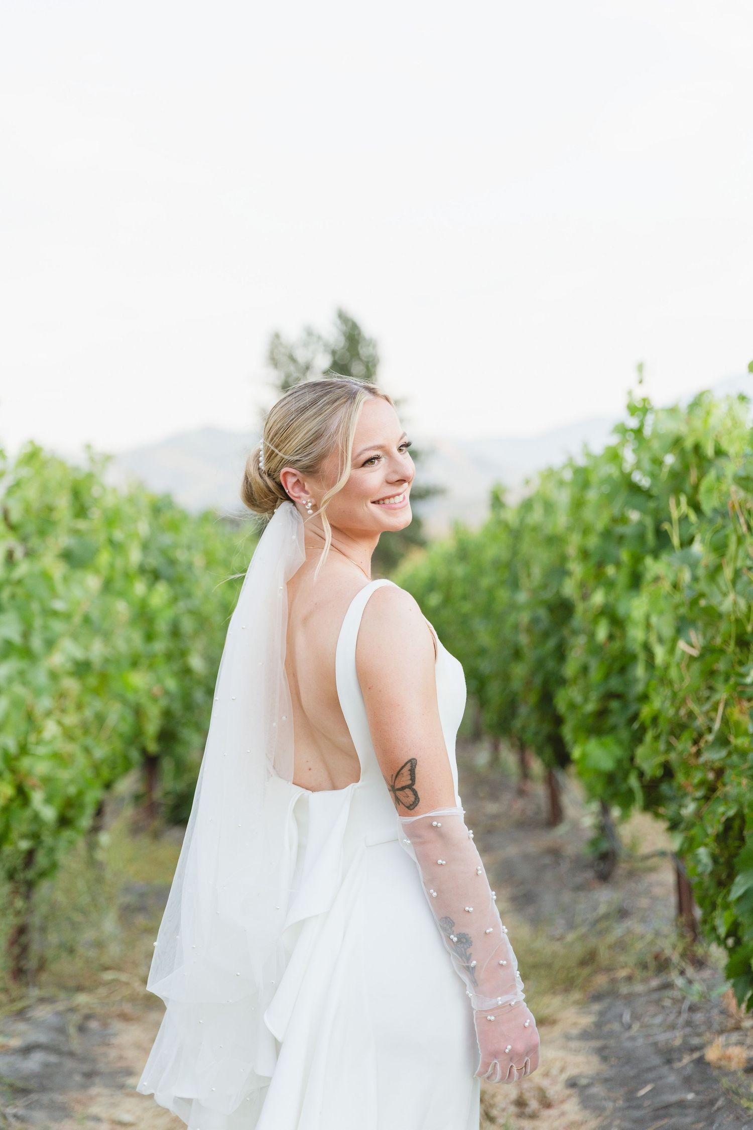 Bride in a backless white wedding dress and veil smiling while standing in a vineyard on a sunny day.
