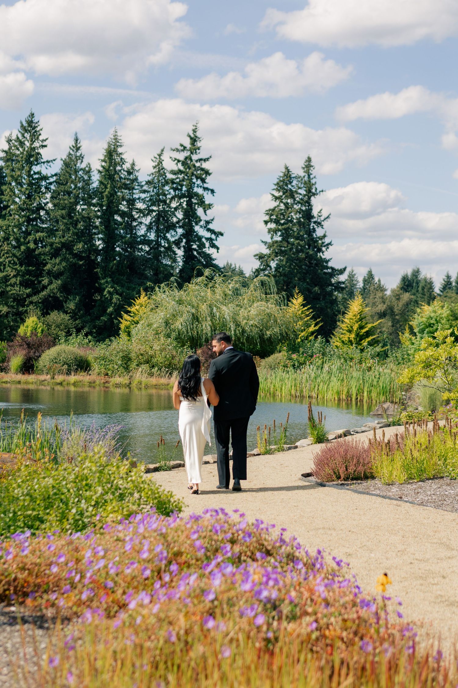 Two people walk along a scenic lakeside path bordered by purple wildflowers and evergreen trees on a sunny day.