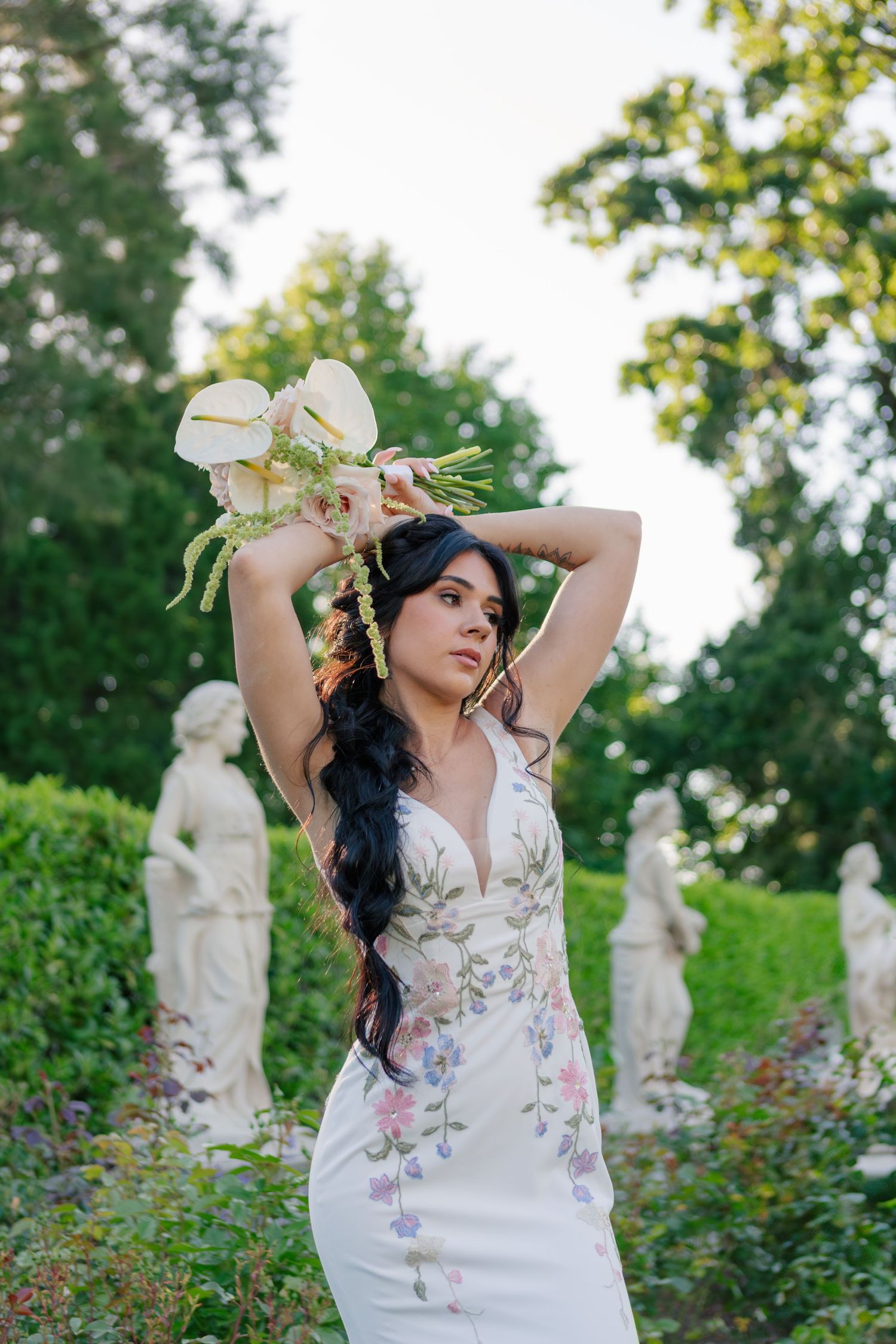 Elegant woman in floral white dress poses gracefully with flower bouquet in garden with classical statues in background.
