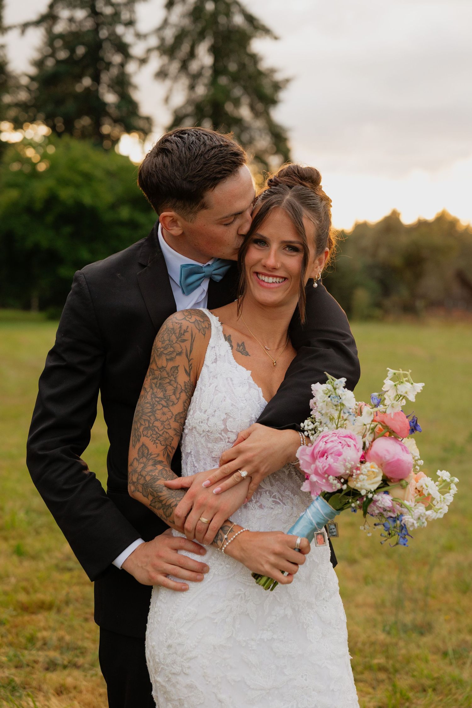 Groom with tattoos kisses bride on cheek as she smiles holding pink floral bouquet in outdoor wedding photo.