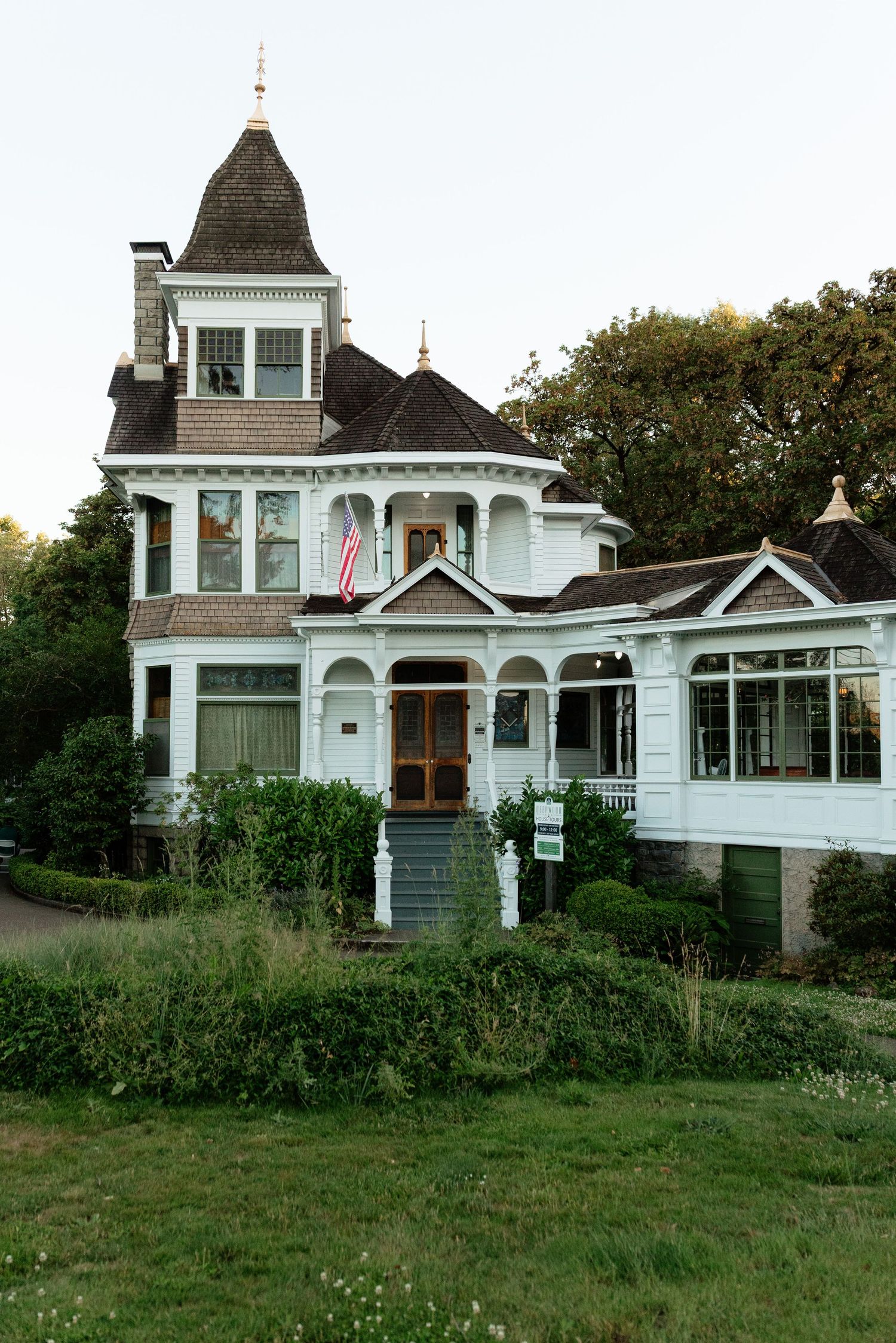 White Victorian mansion with dark turret roof, American flag, wrap-around porch, and lush green garden landscaping.