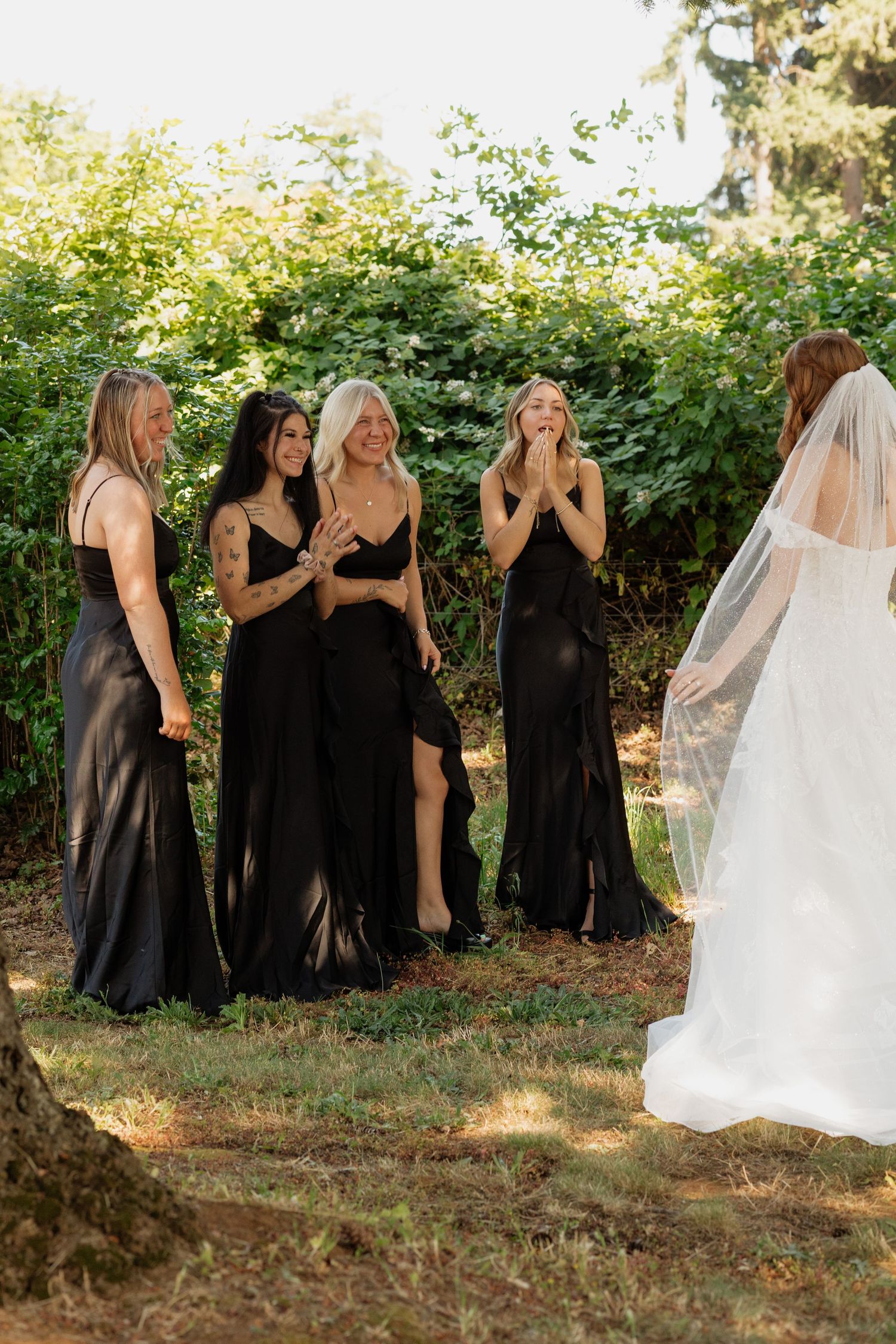 Three bridesmaids in black dresses react with joy seeing the bride in her white wedding gown outdoors.