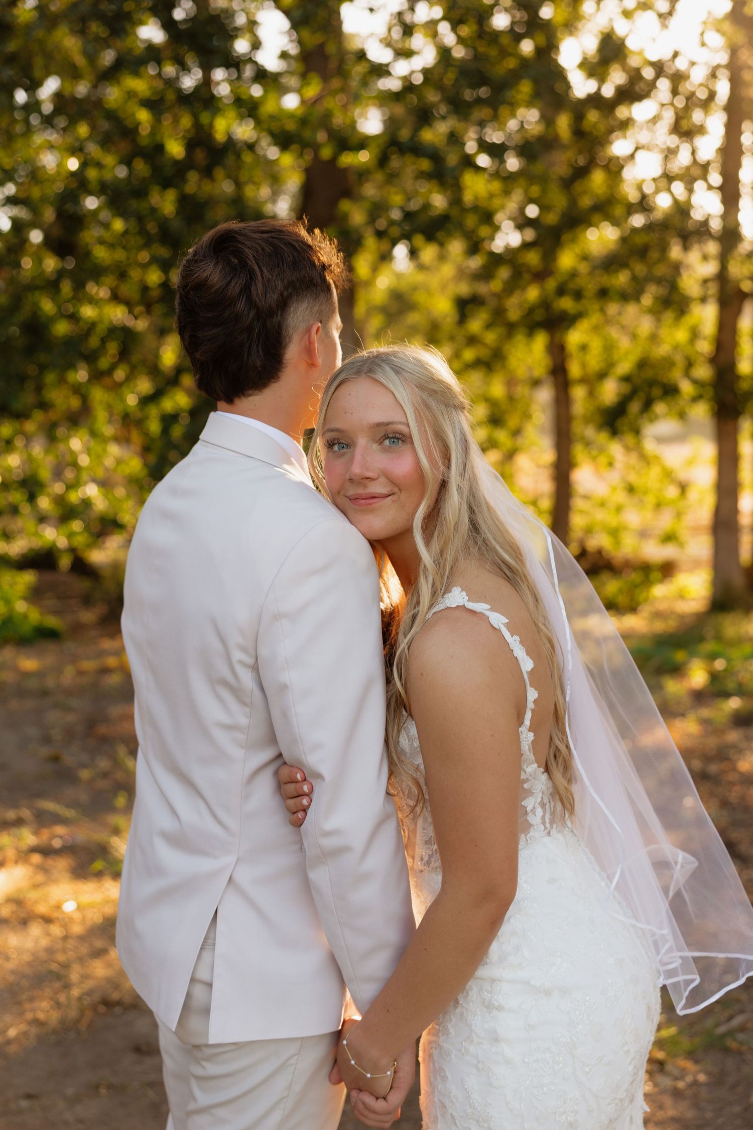 Bride in lace gown glances back at camera while holding hands with groom at golden hour.