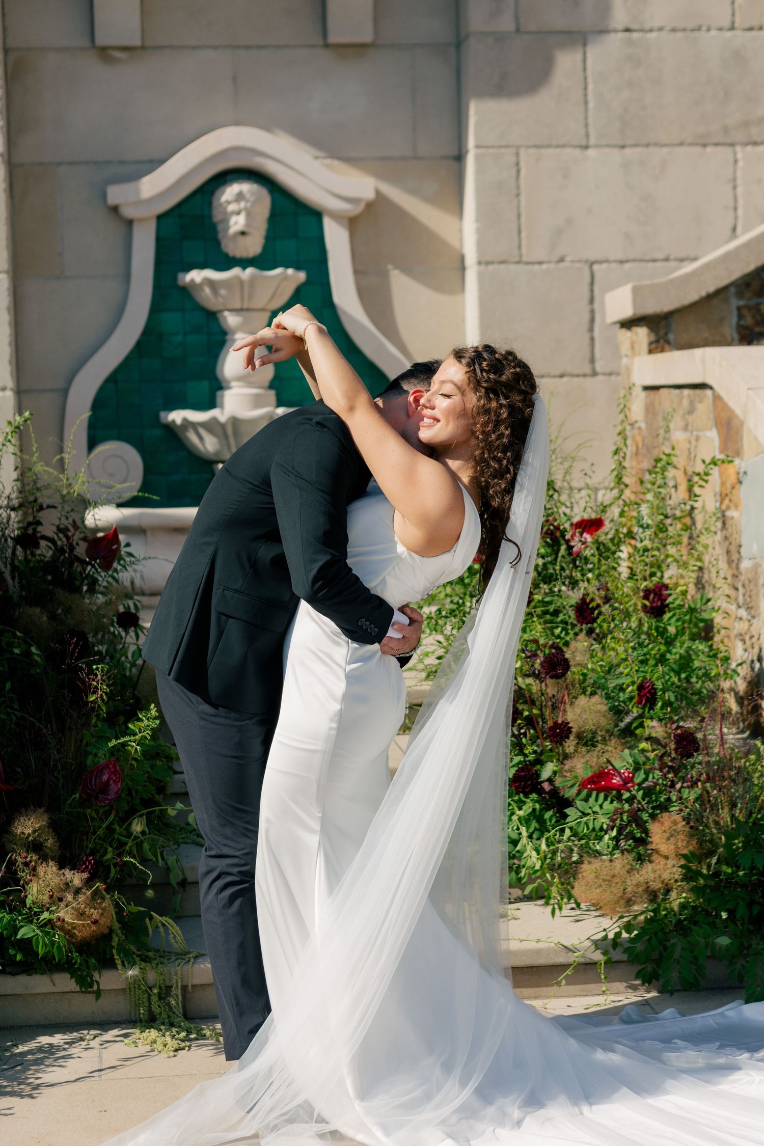 A joyful wedding couple embraces in a romantic dip pose against a garden backdrop with a fountain and blooming flowers.