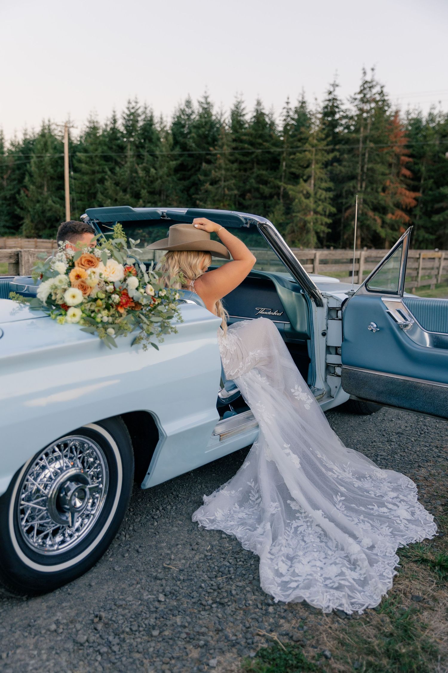 Bride in lace gown and cowboy hat leaning into vintage light blue convertible car holding floral bouquet outdoors.