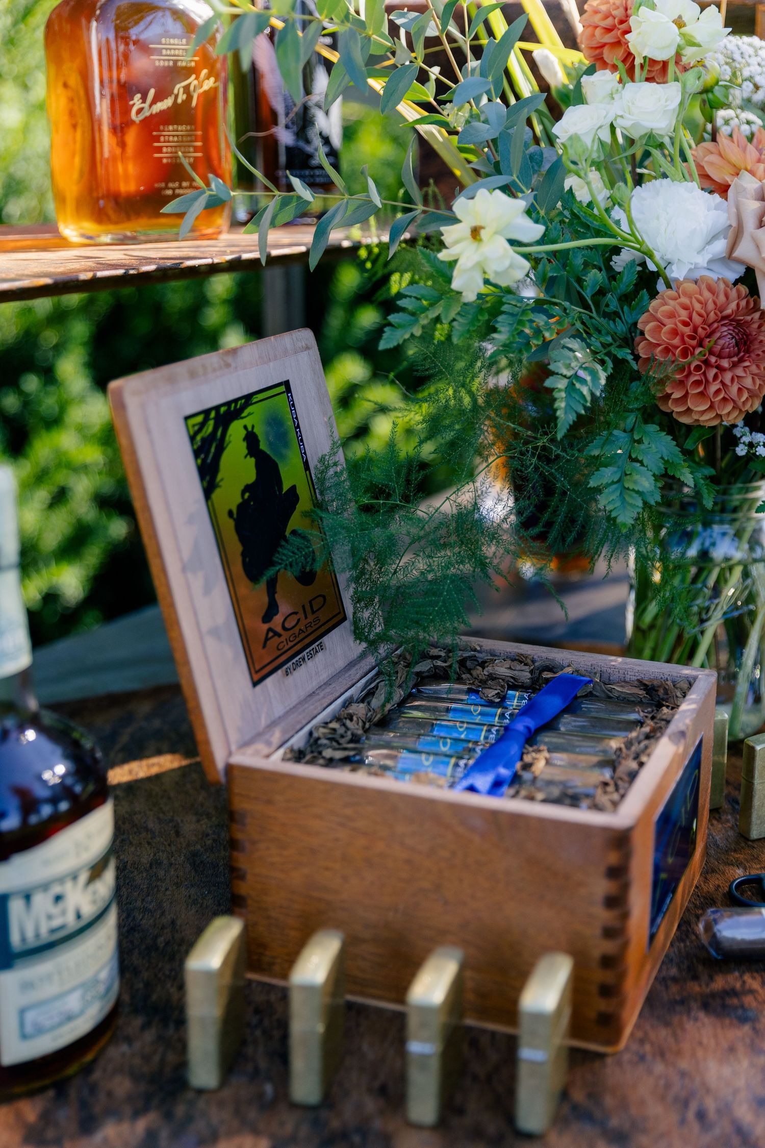 Wooden cigar box with cigars displayed on a rustic bar setup with whiskey bottles and floral arrangements at a wedding.