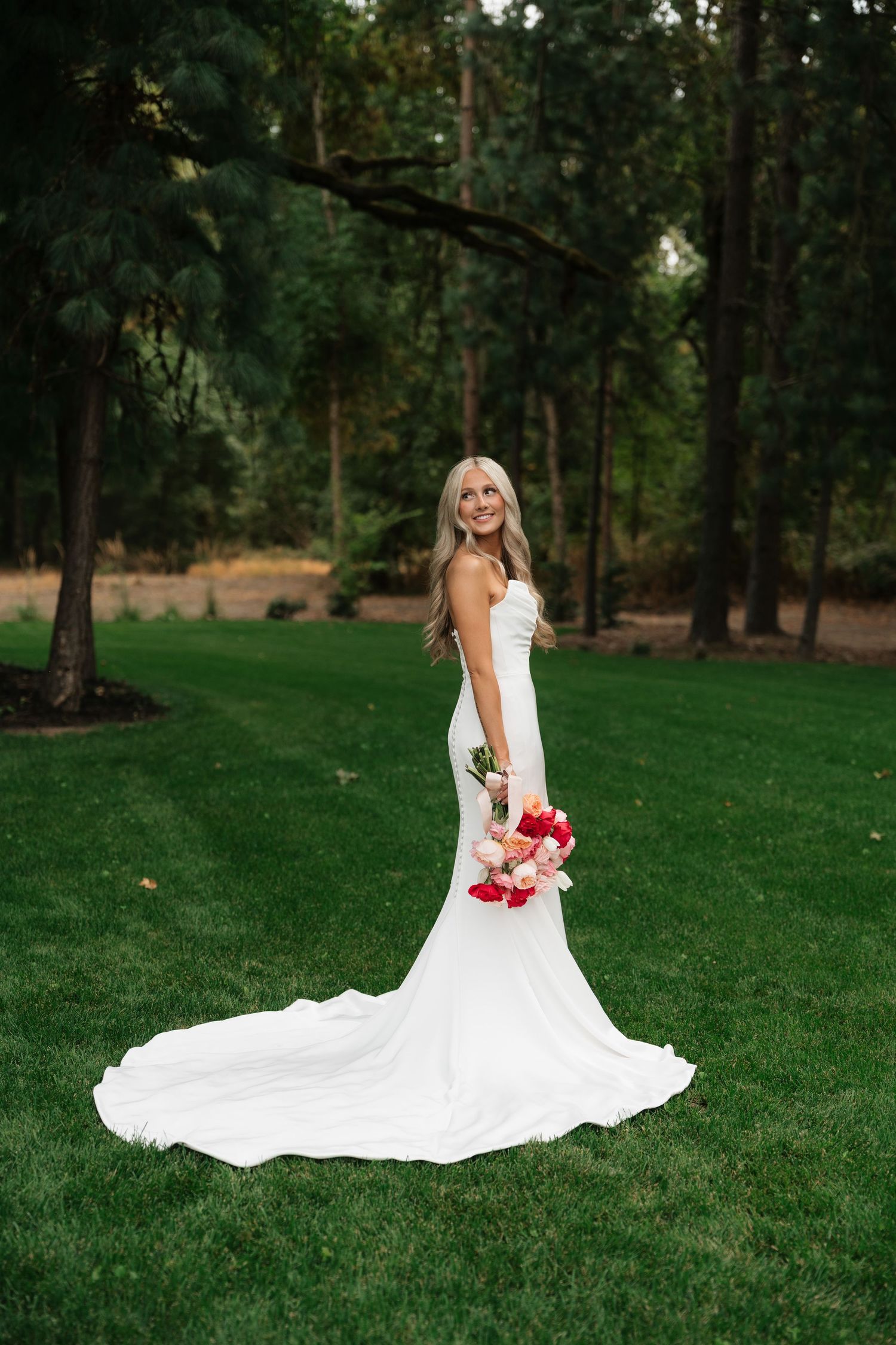 Bride in white mermaid gown with train holds red bouquet on green lawn surrounded by tall pine trees.